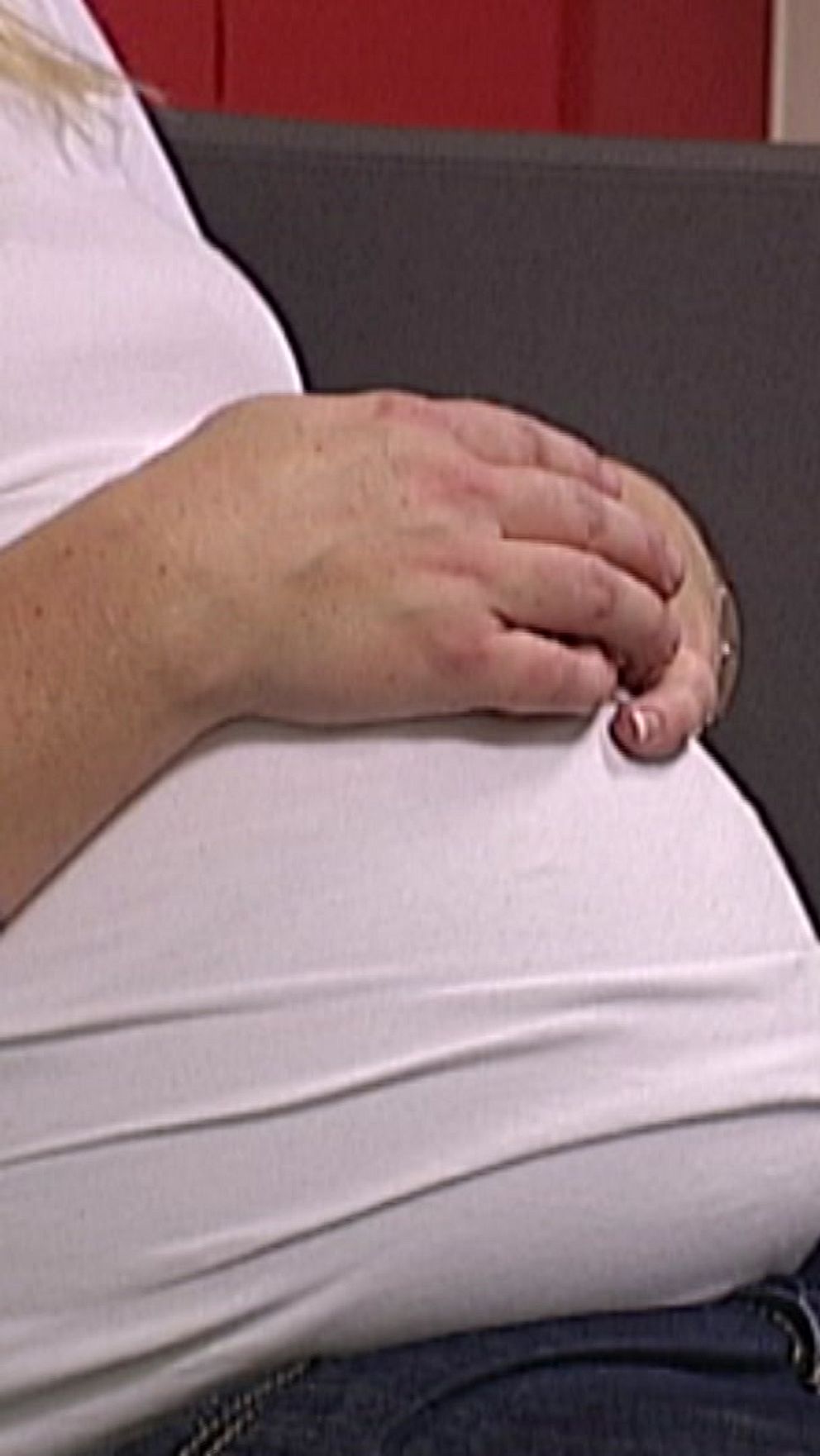 Gravid kvinna i vit tröja sitter på en soffa, håller händerna på magen.