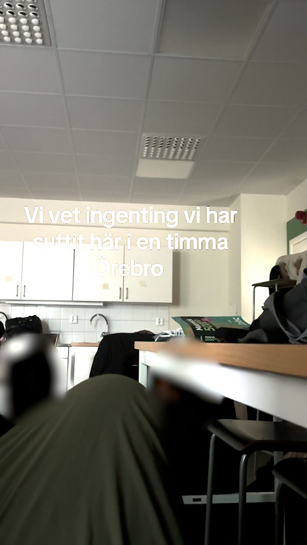 En person sitter i ett klassrum med vita skåp och bänkar. Texten över bilden lyder: ”Vi vet ingenting vi har suttit här i en timma Örebro”.