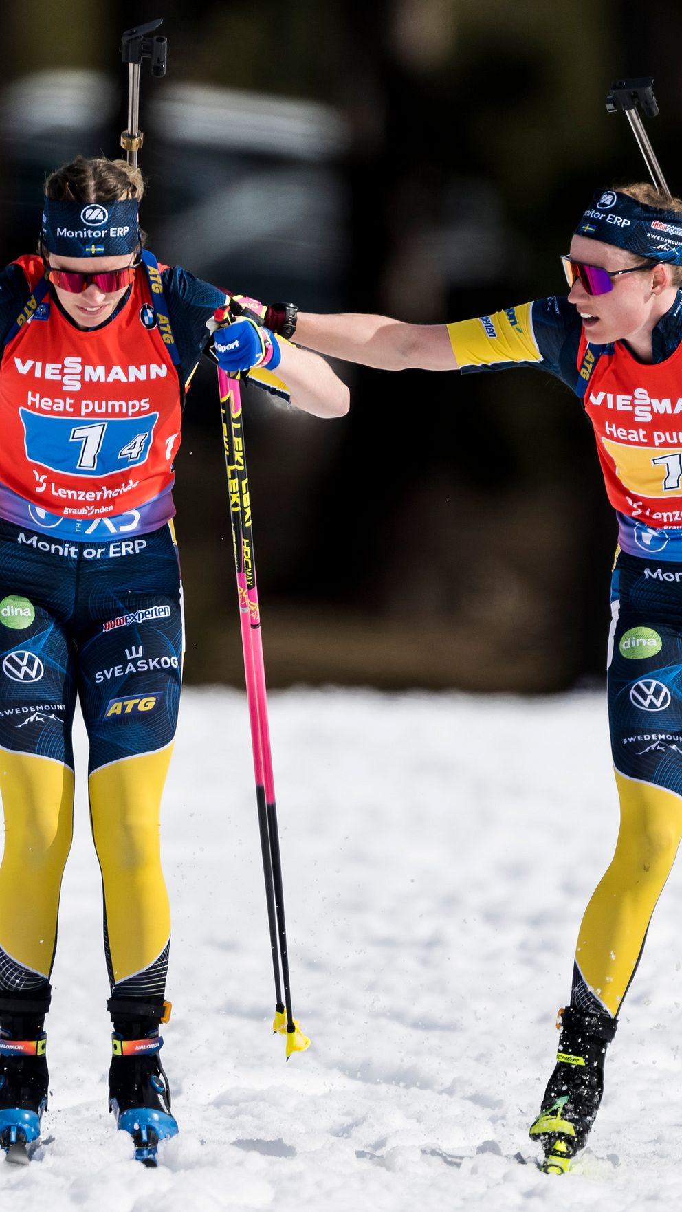 Två svenska skidskyttar i tävlingsdräkt hjälper varandra på snötäckt bana.