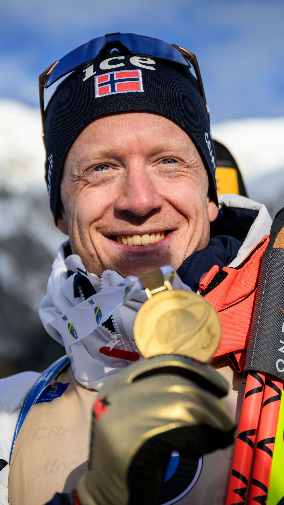 En skidåkare iförd hjälm och skidutrustning håller upp en guldmedalj framför ett snöklätt bergslandskap, med ett par Fischer-skidor i ena handen.