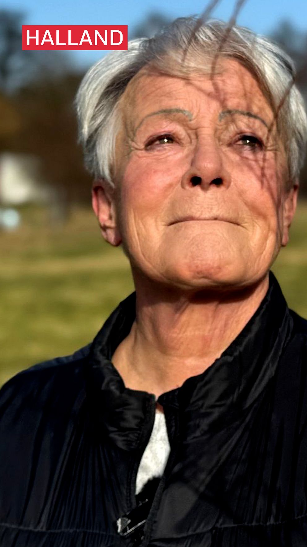Person i mörk jacka står utomhus med grönt fält i bakgrunden.