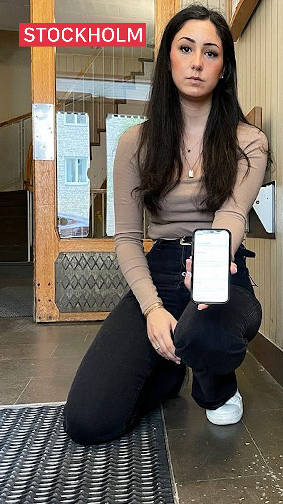 Kvinna håller fram mobil och sitter vid ett hål i dörren, som orsakats av en sprängning.