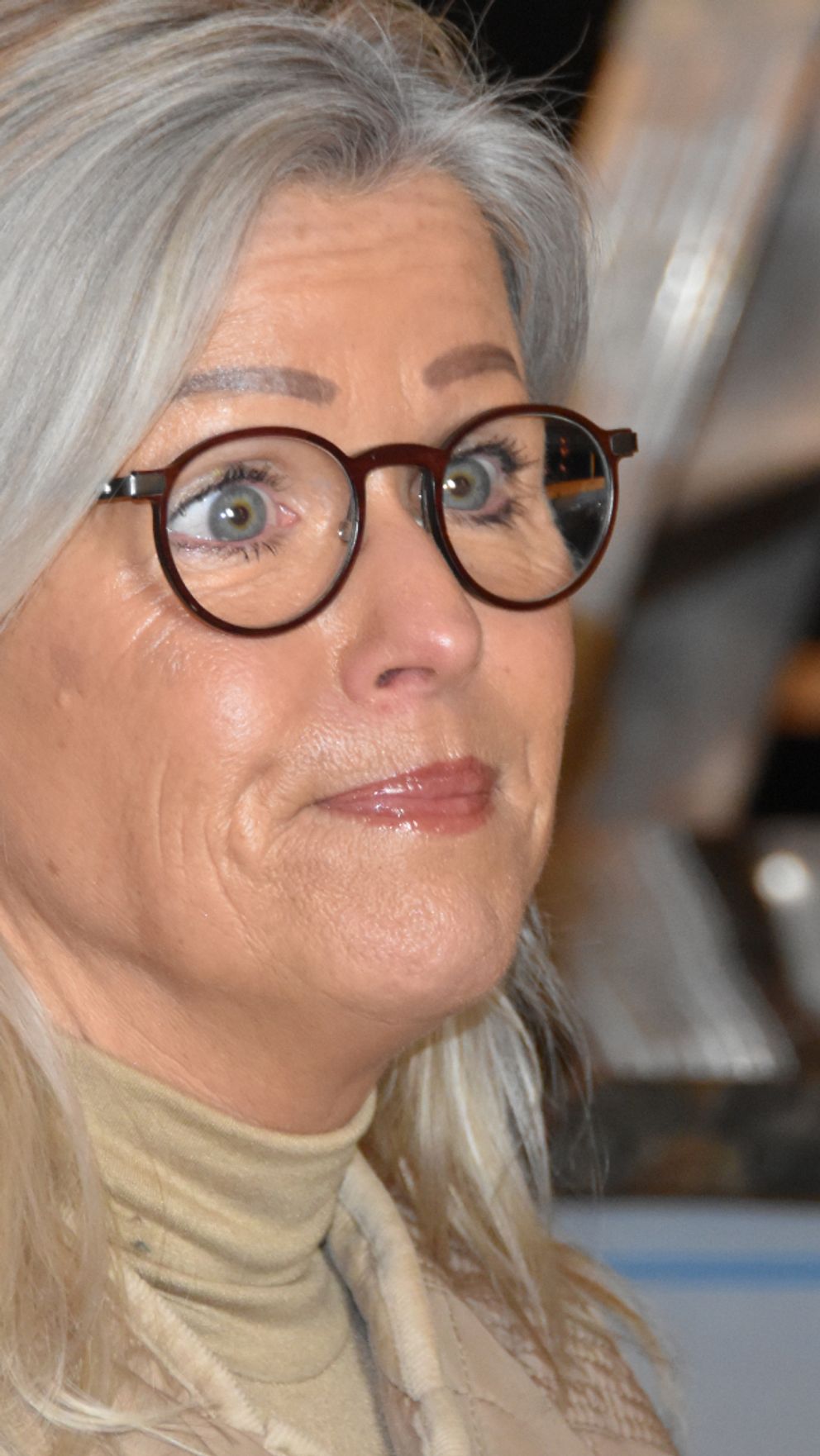Åsa Wiklund Lång (S) med långt, grått hår som bär en beige tröja och örhängen, står i en inomhusmiljö med en stege i bakgrunden.
