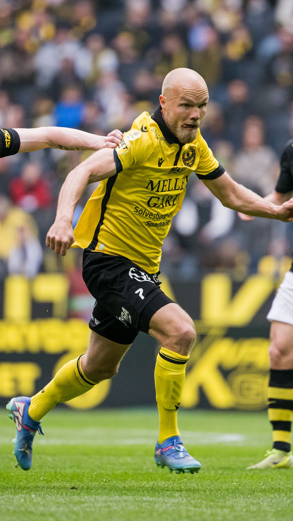 Fotbollsmatch: tre spelare jagar bollen, två i svart och en i gul matchtröja.