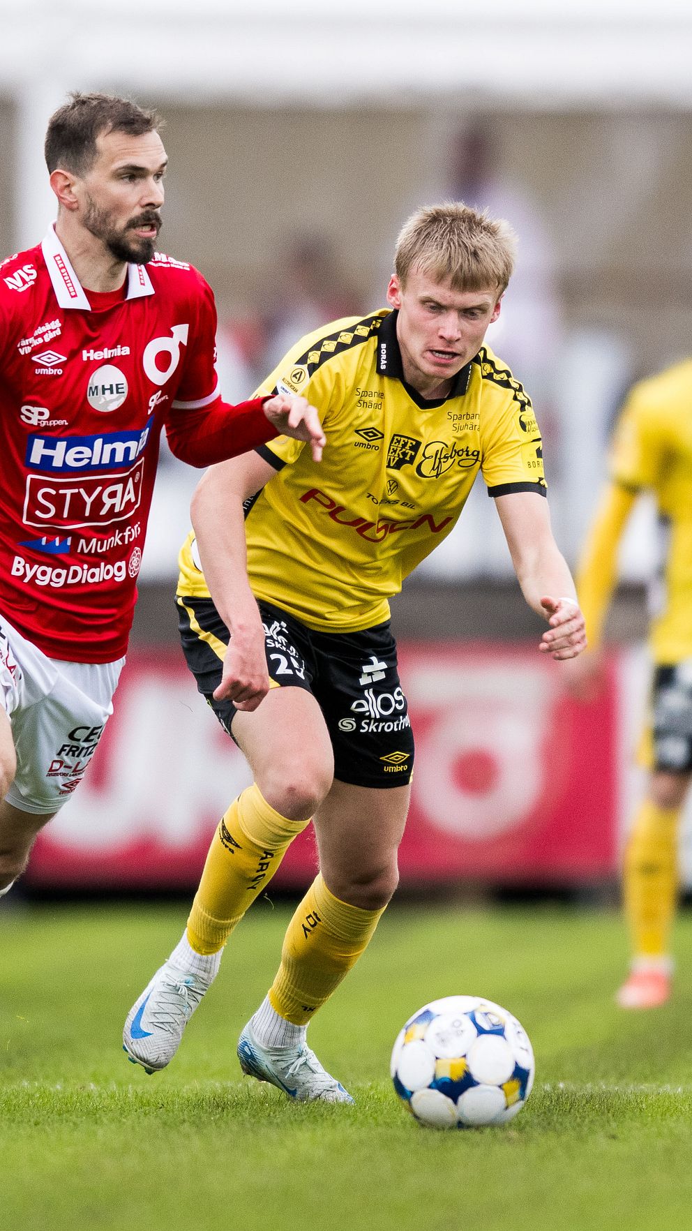 Fotbollsspelare i röd och gul tröja jagar boll på planen under match.