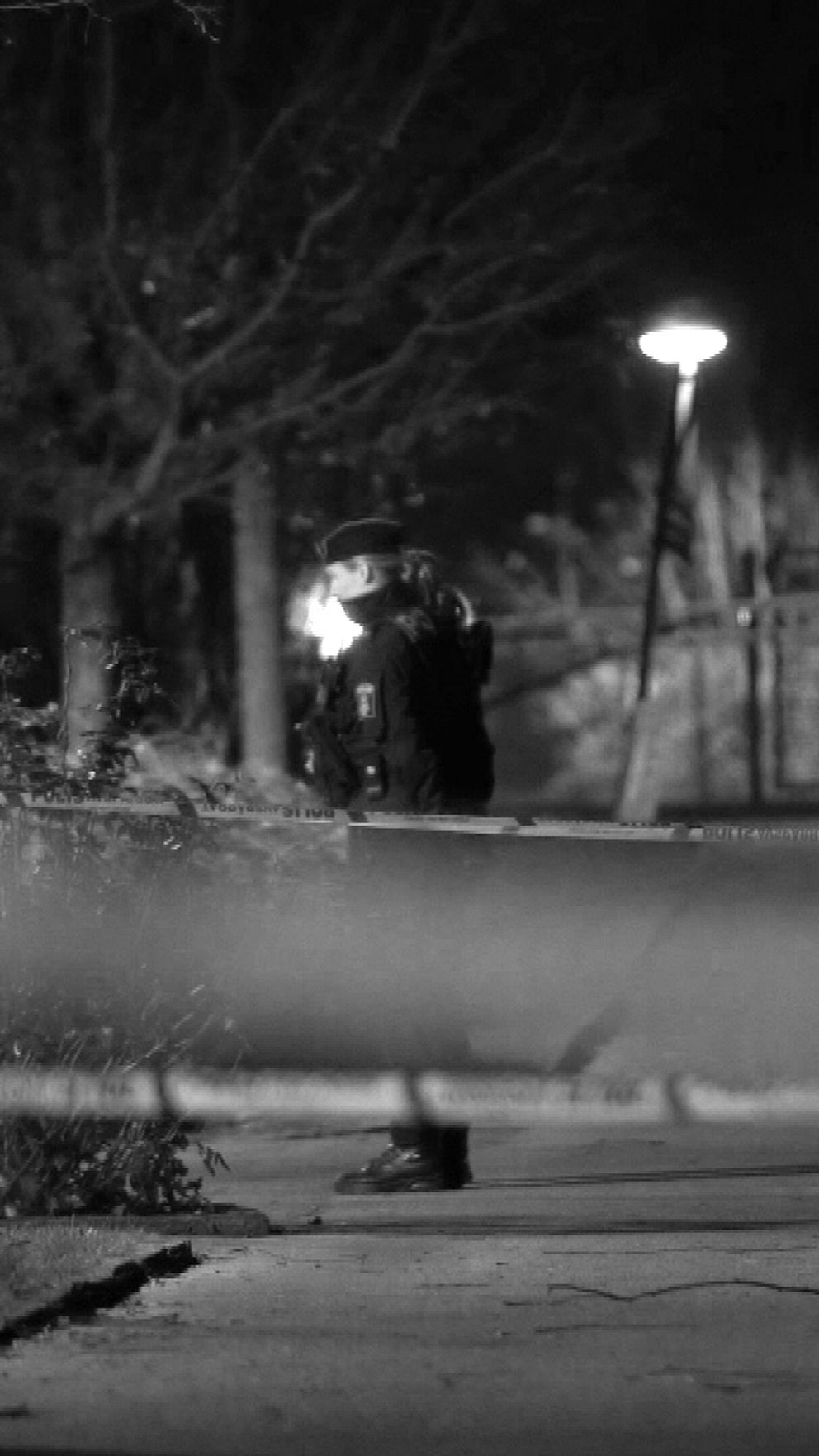 Poliser arbetar vid en avspärrning i en dämpad belysning under nattens mörker, med fokus på att utföra en undersökning på en plats som tyder på en incident.