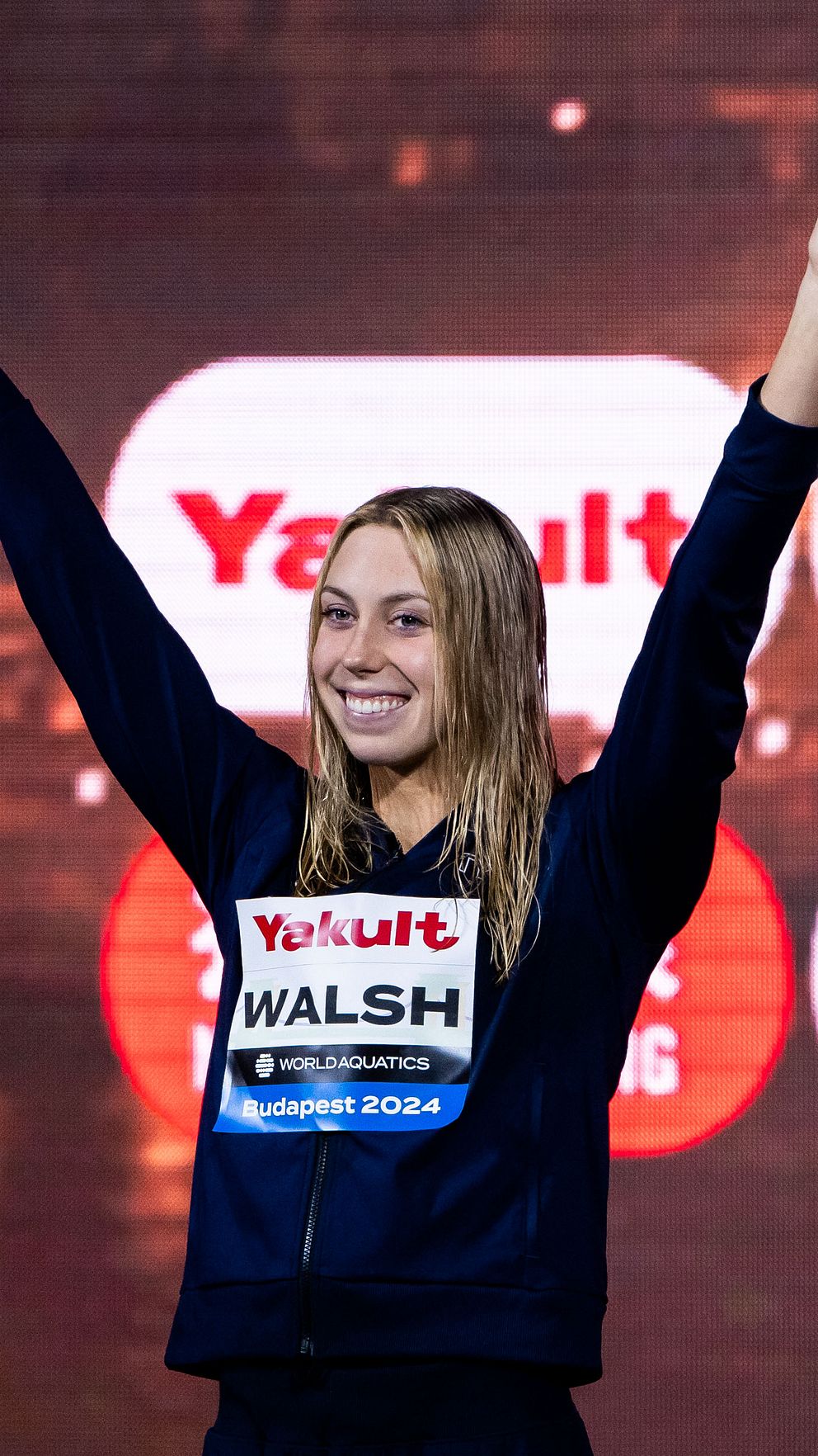 En idrottare sträcker upp sina armar i luften som en gest av seger eller glädje, iförd en mörk outfit med sponsorlogotyper, inklusive ”Yogurt” och ”Walsh.” Bakgrunden visar suddiga ljus och logotyper för företag relaterade till evenemanget.