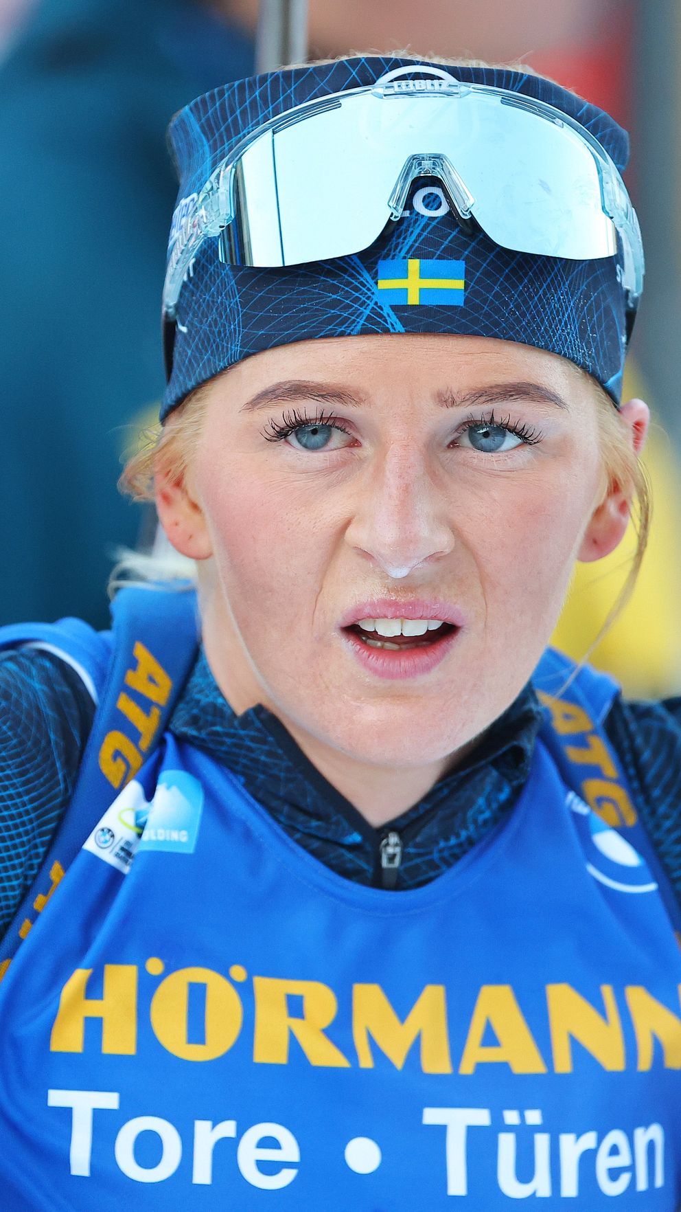 En skidskytt står med armarna utsträckta och har på sig en tävlingsdräkt i blått och gult med sponsorns namn ”HÖRMANN Tore + Türen” på bröstet. Bakgrunden är suddig men antyder en sportevenemangsmiljö. Skidskytten har också skyddsglasögon på huvudet.