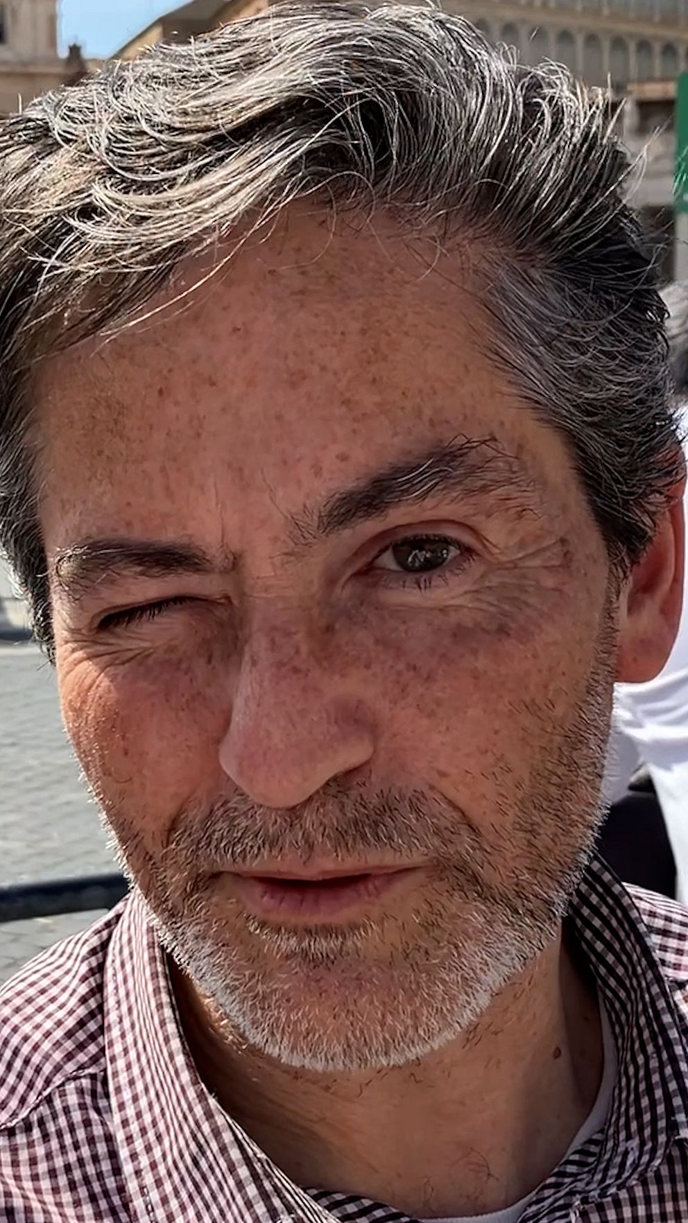 Person med grått hår i solsken. Bakom syns historisk byggnad.
