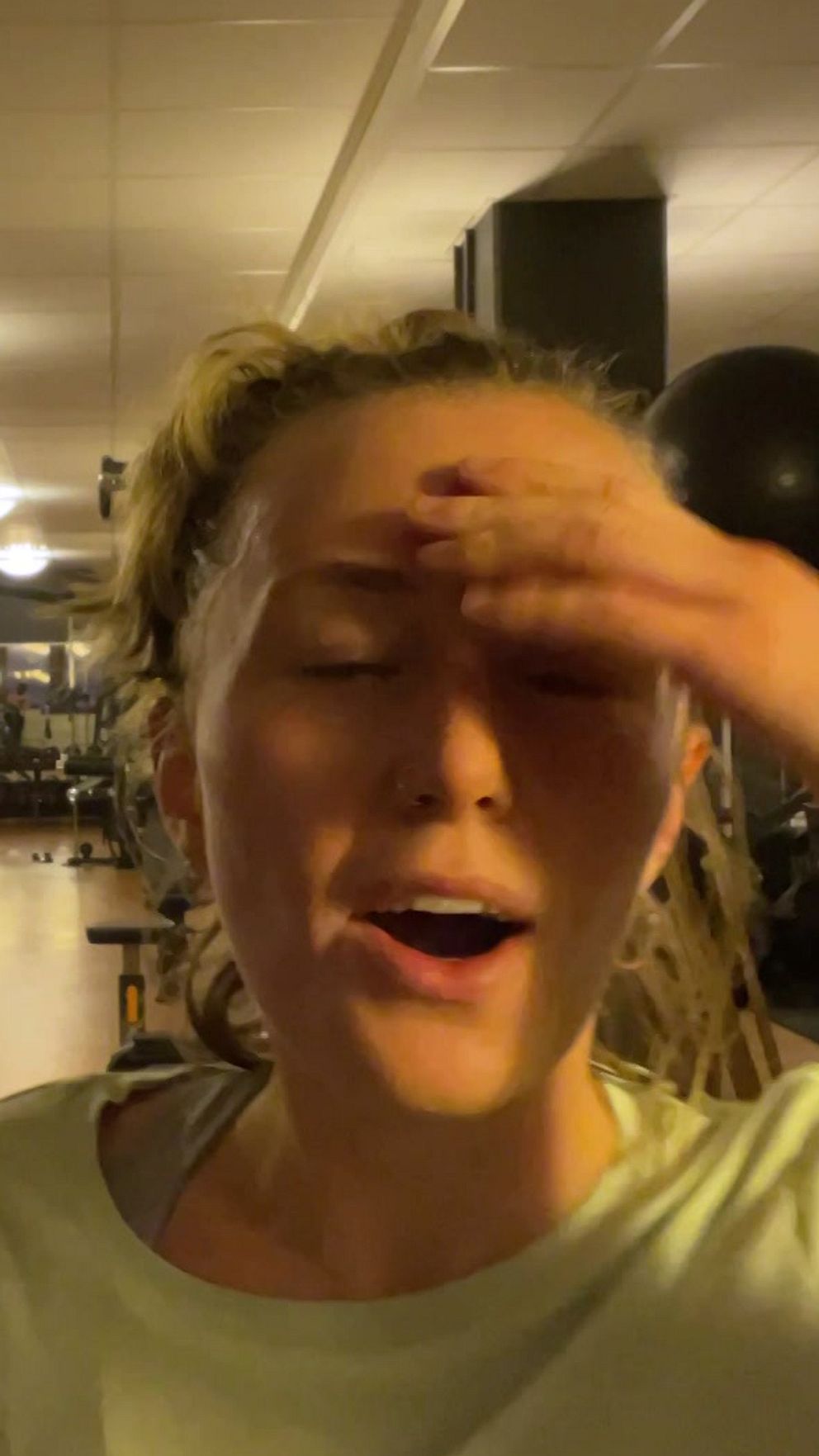 En person står i ett gym med träningsutrustning, som viktmaskiner och bänkar, i bakgrunden. Gymmet är upplyst med taklampor och har speglar längs ena väggen.