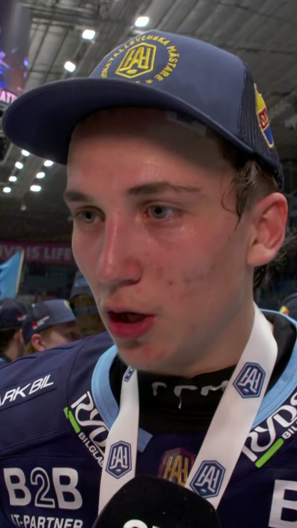 Hockeyspelare med guldmedalj intervjuas efter match, andra lagkamrater syns i bakgrunden.