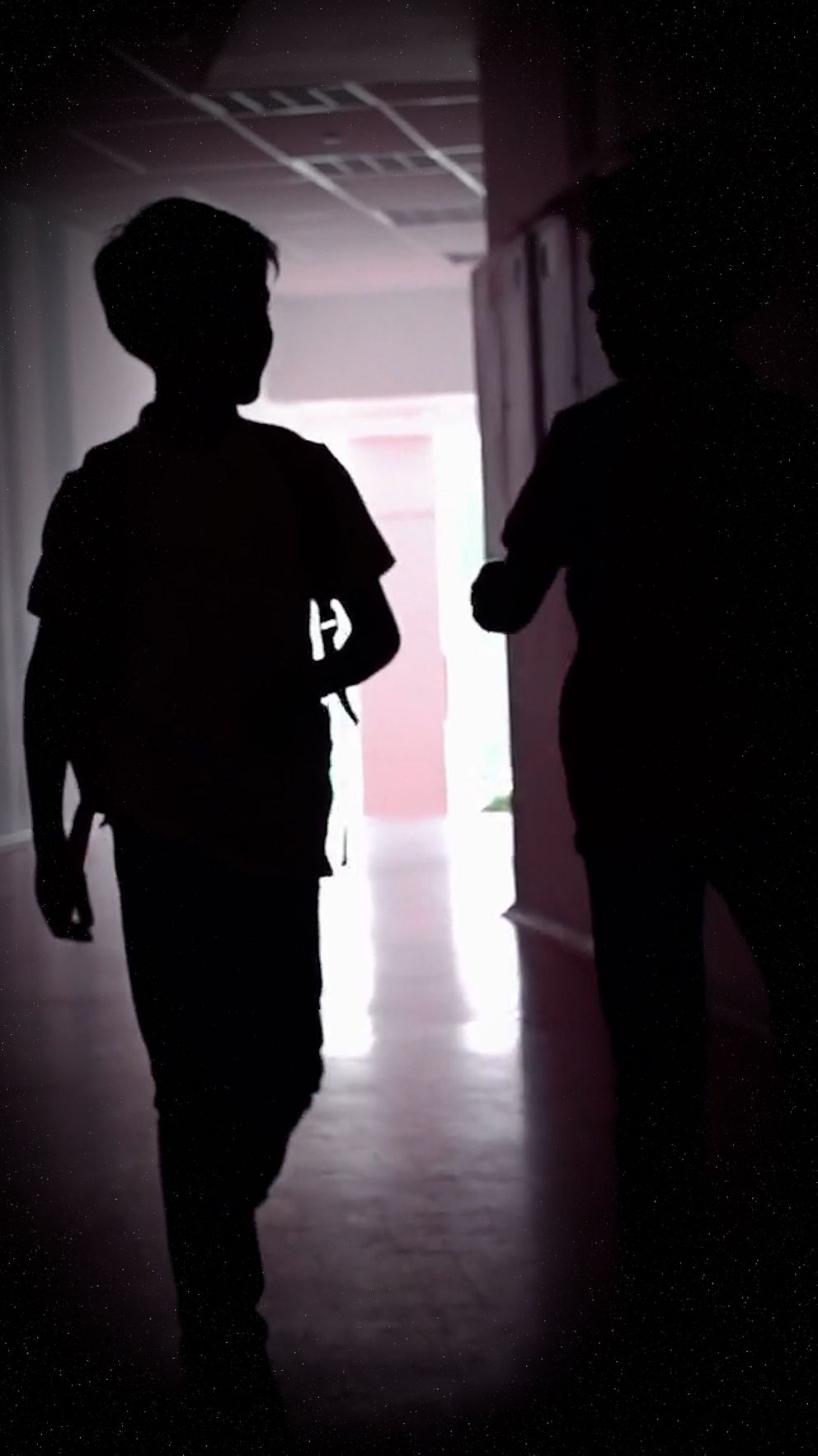 Två silhuetter av barn som går i en korridor, med svagt ljus som strömmar in från en dörr i bakgrunden. Den ena personen har en ryggsäck, medan den andra verkar motionera med en hand i rörelse. Korridoren har en modern känsla med en ljus och öppen atmosfär.