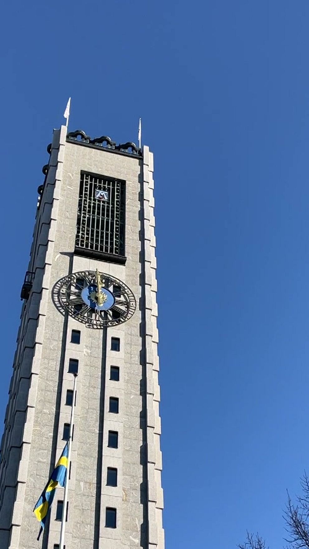 Högt klocktorn mot en klarblå himmel, med svenska flaggor som vajar i vinden och trädgrenar synliga i högra sidan av bilden.