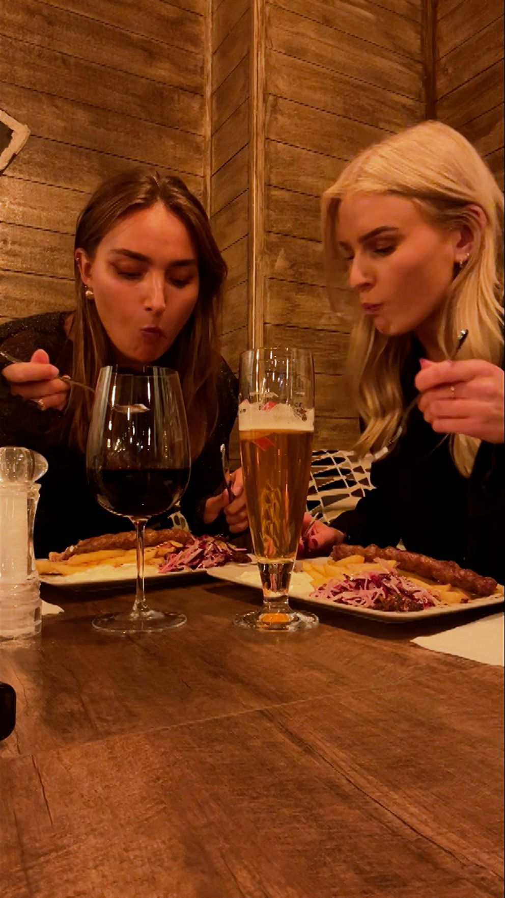 Två personer sitter vid ett middagsbord, omgivna av en trävägg. På bordet finns två tallrikar med mat, samt ett glas vin och ett ölglas. De verkar njuta av en måltid tillsammans. Det är en varm och mysig atmosfär i rummet.