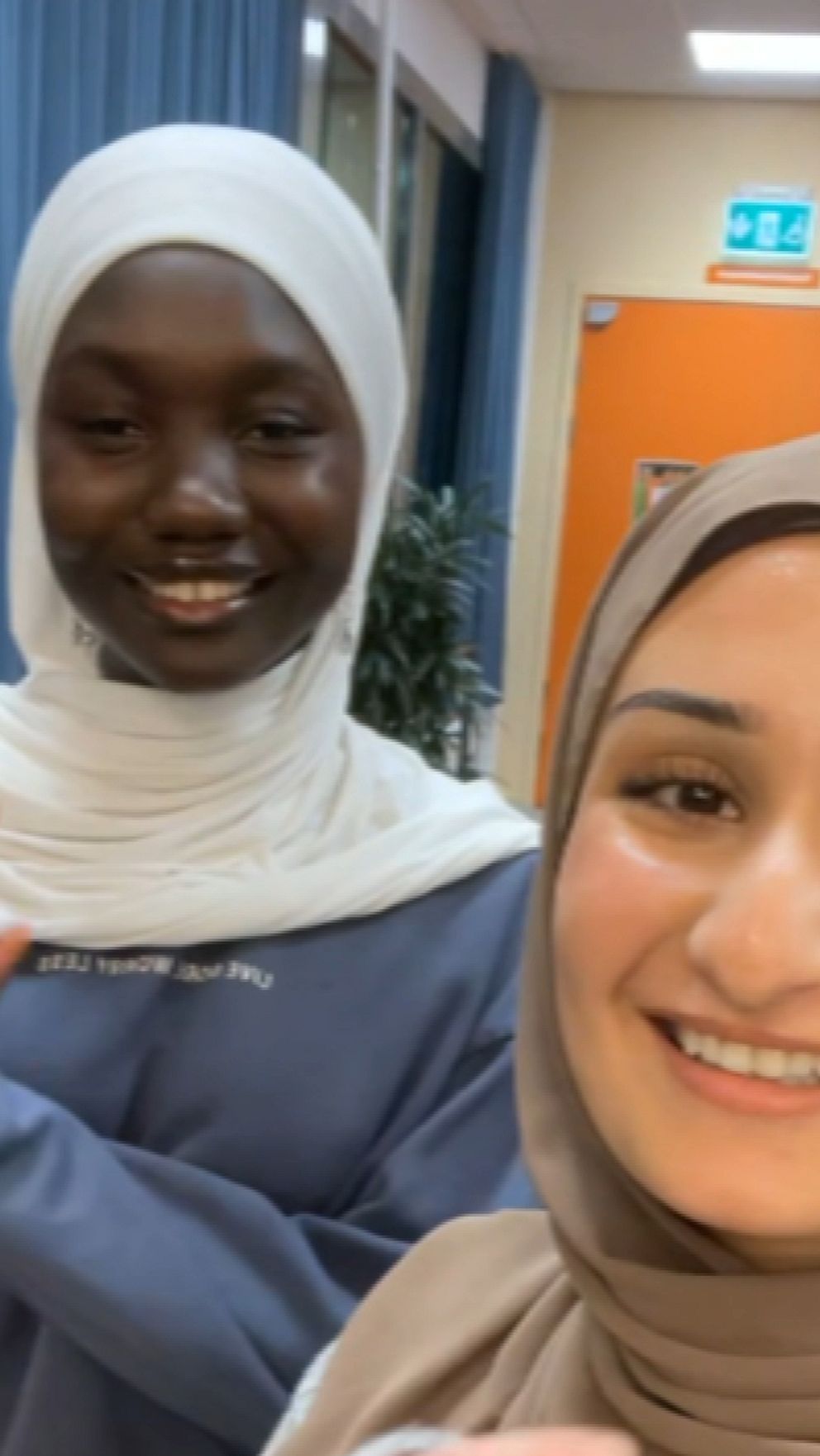 Två personer står i en inomhusmiljö, båda klädda i hijab. Den första personen, till vänster, har på sig en ljusblå tröja och gestikulerar med handen. Den andra personen, till höger, bär en beige hijab och ler. Bakgrunden innehåller inredning med blå väggar och fönster som släpper in ljus.
