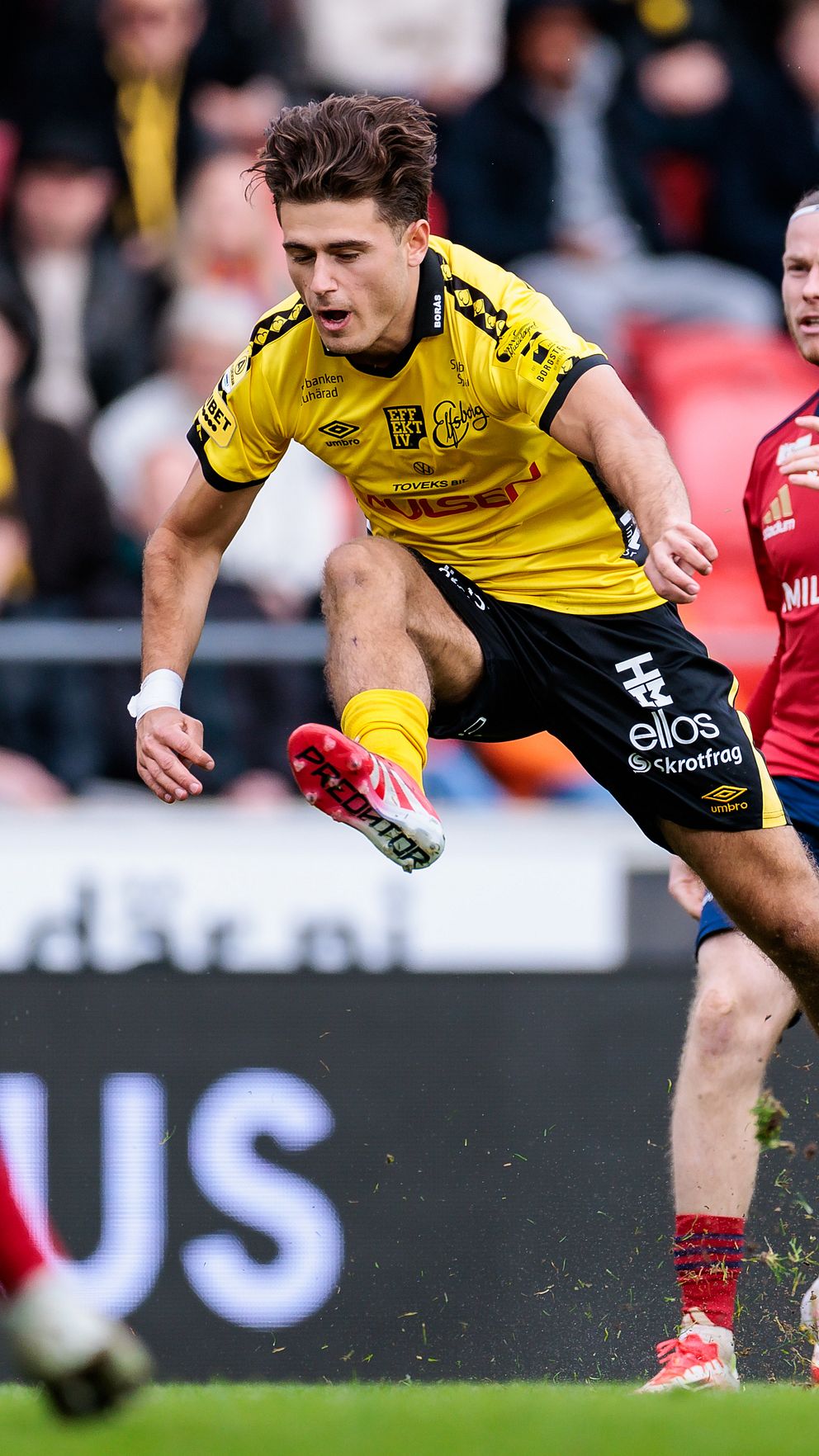 Fotbollsspelare i gul tröja hoppar för att sparka bollen mot motståndare i röda tröjor.