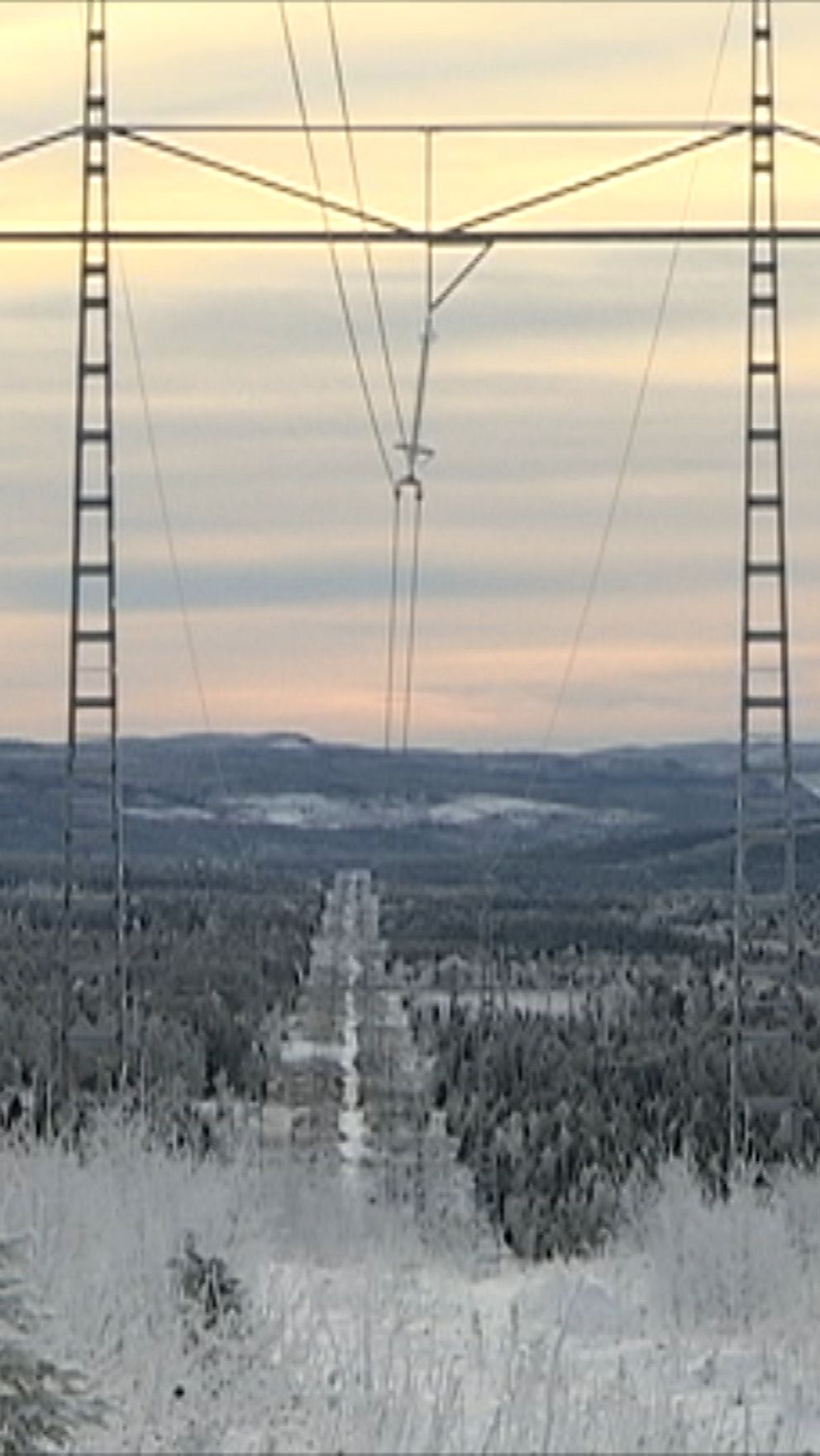 Snöklädda träd och en vidsträckt dalgång avbildas under en gryningshimmel med mjuka pastellfärger. Stora kraftledningsmaster reser sig i förgrunden, omgivna av en fryst landskap som sträcker sig över horisonten.