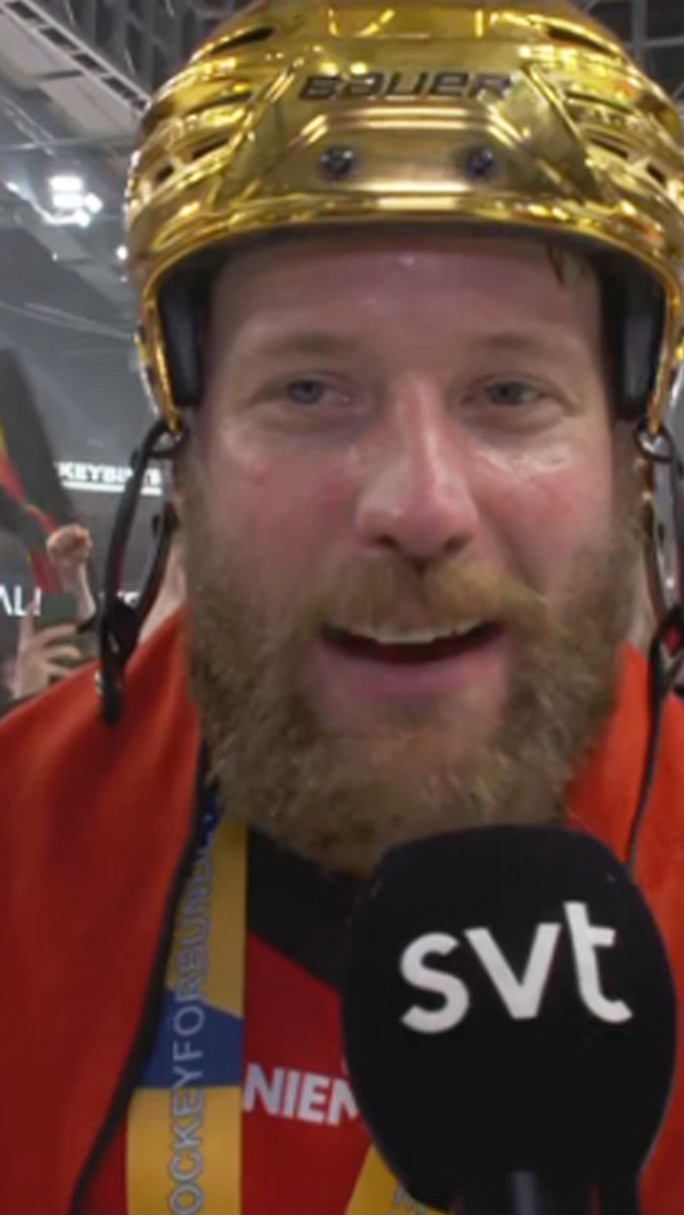 Hockeystjärna med guldhjälm intervjuas på isen, fans firar i bakgrunden.