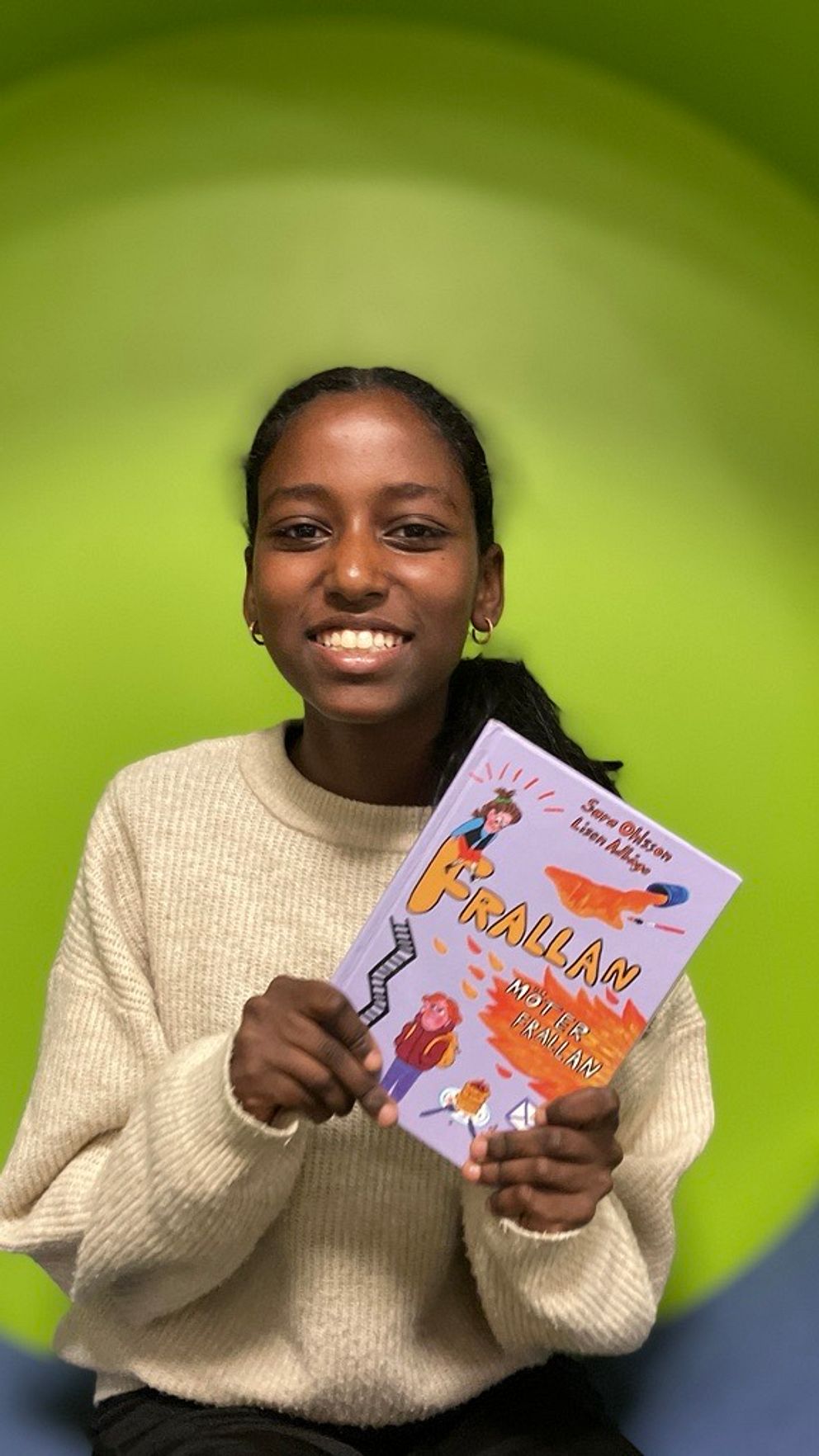 En person håller upp en bok med titeln ”Frallan” framför en grön bakgrund. Personen är klädd i en ljus, stickad tröja och ser ut att vara glad eller entusiastisk över boken.