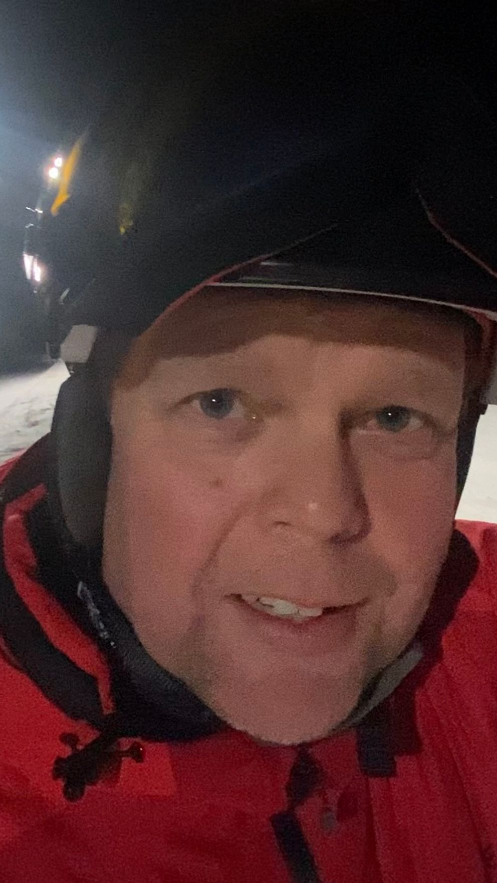 En person i en röd skidjacka står på en snötäckt skidbacke under natten, omgiven av belysning från lampor längs backen.