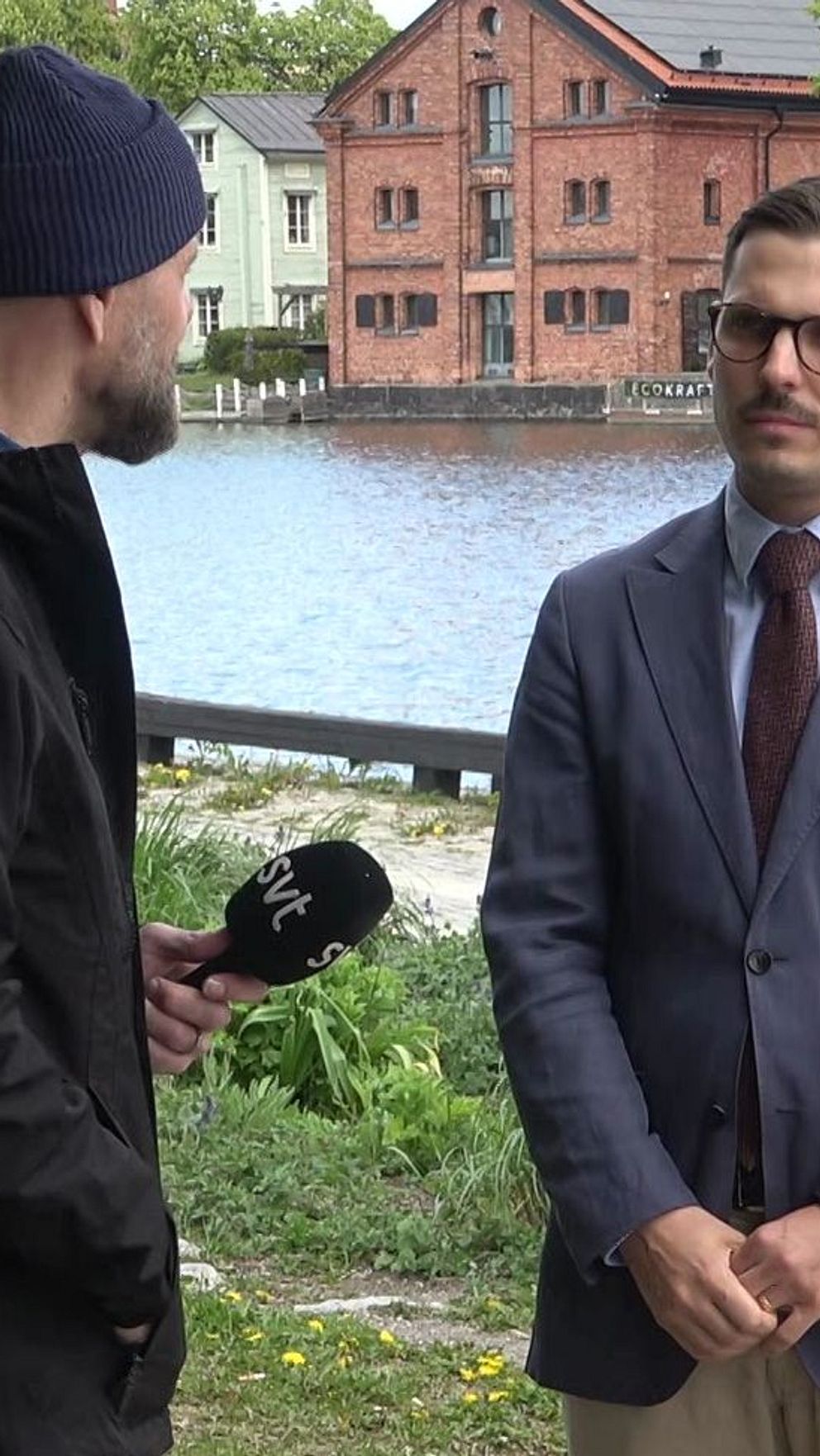 Reporter intervjuar man i kostym vid vattnet med byggnader i bakgrunden.