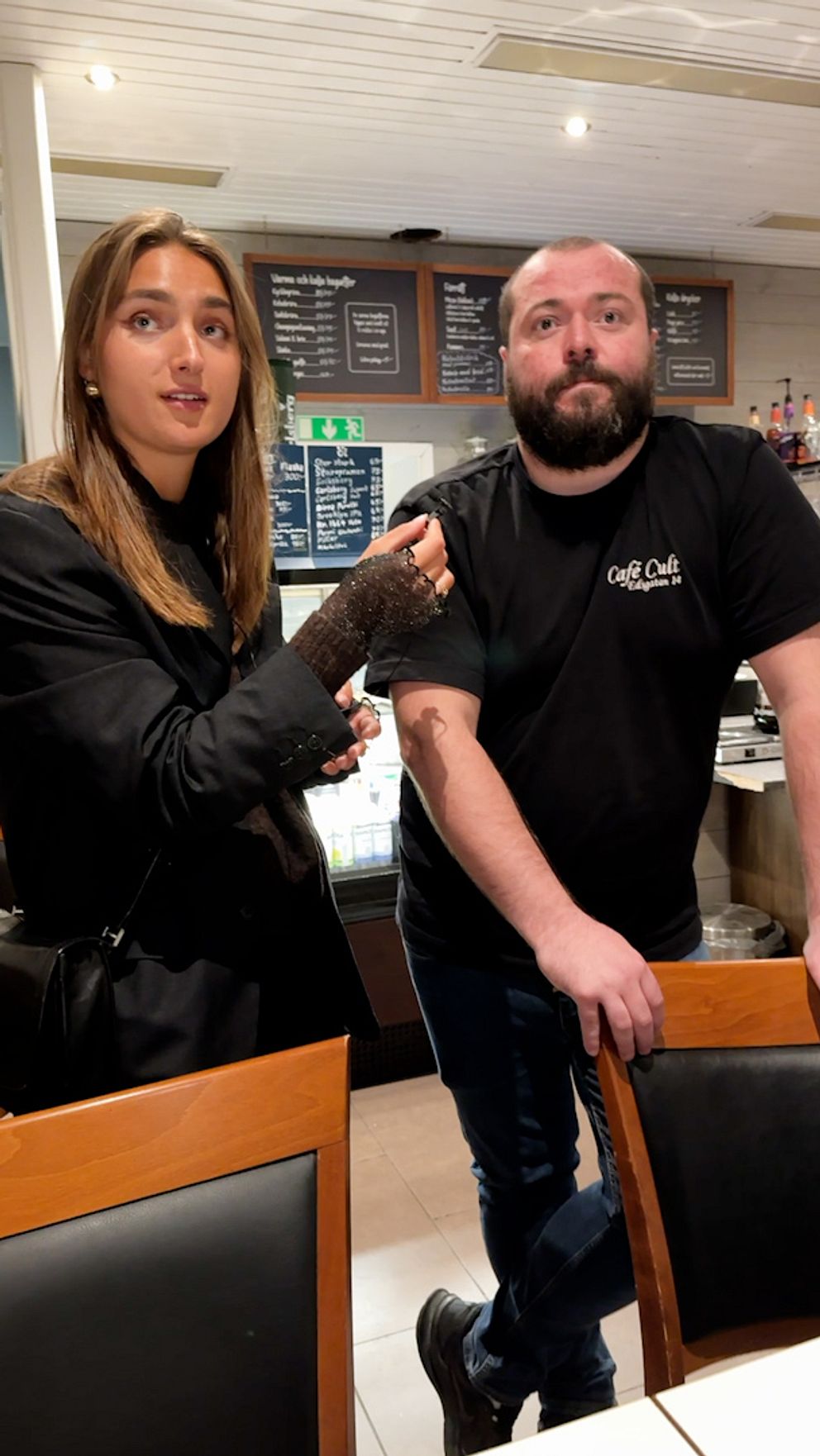 En kvinna står i en restaurangmiljö och intervjuar en man som lutar sig mot ett bord. De verkar ha en konversation, omgivna av restaurangens inredning och menyer i bakgrunden.