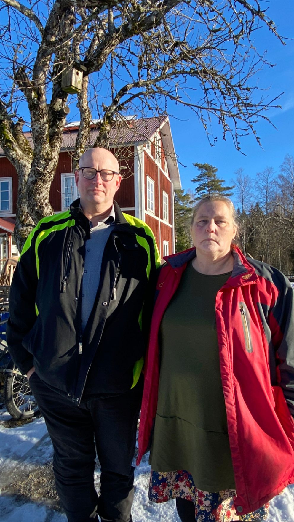 Man och kvinna står framför röd stuga i vintrigt landskap.