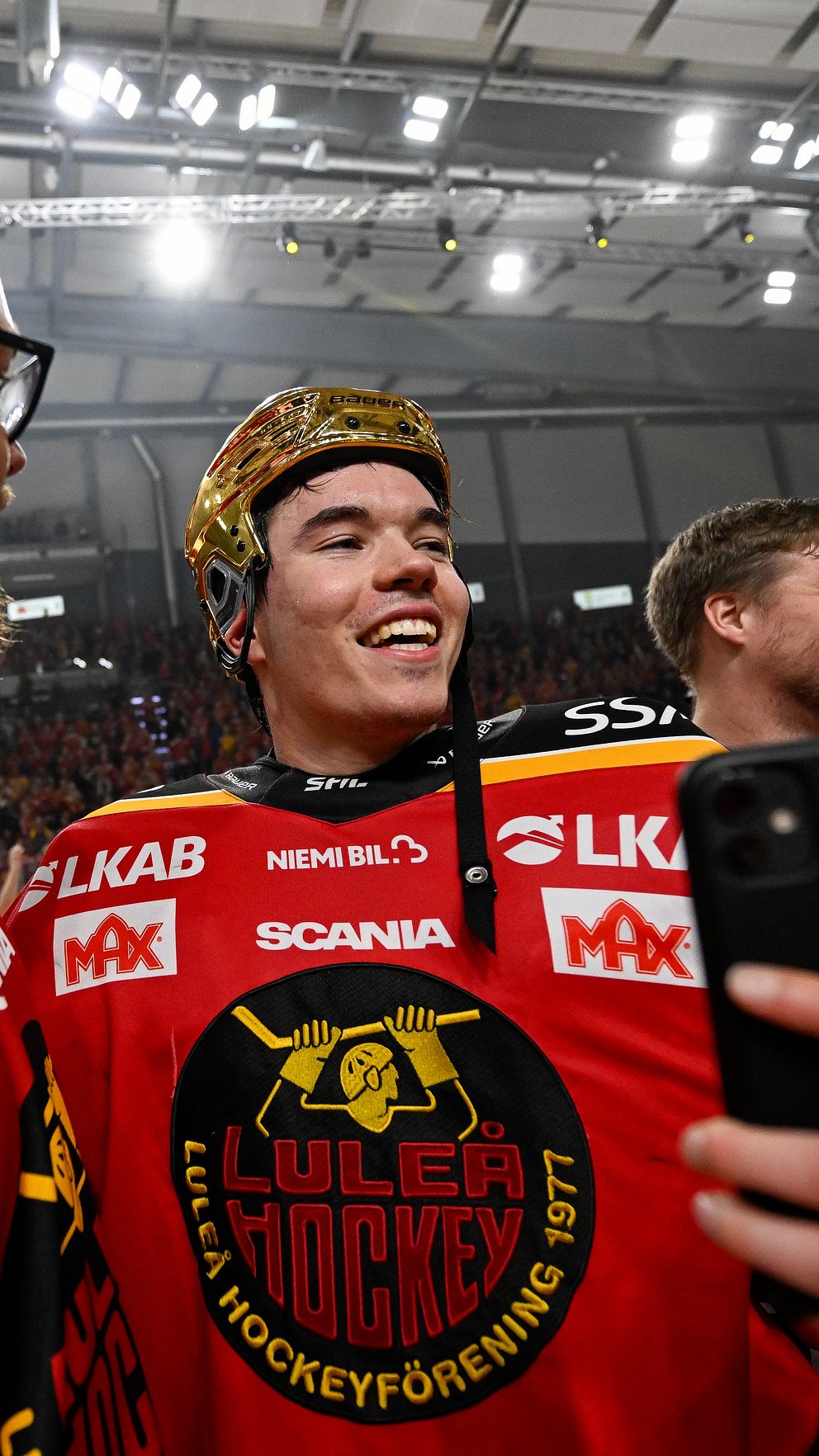 Ishockeyspelare i röda matchtröjor firar på isen, någon tar ett foto med mobil.