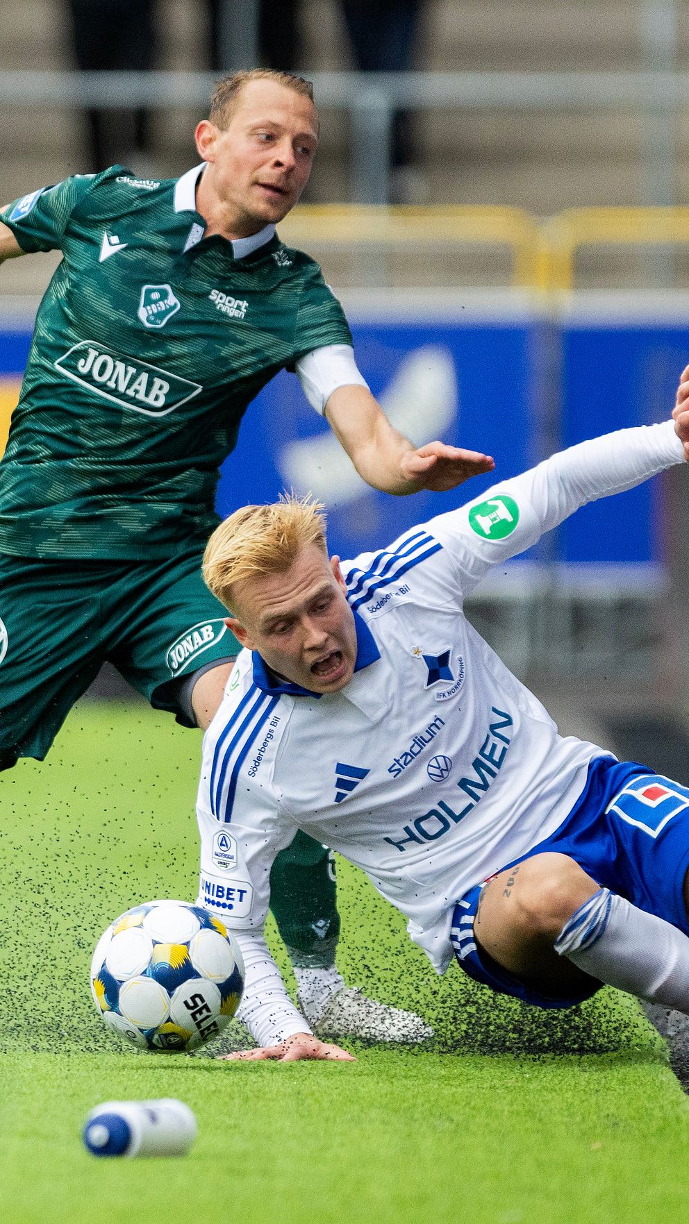Fotbollsspelare kämpar nära sidlinjen under matchen. En spelare faller mot barriären.