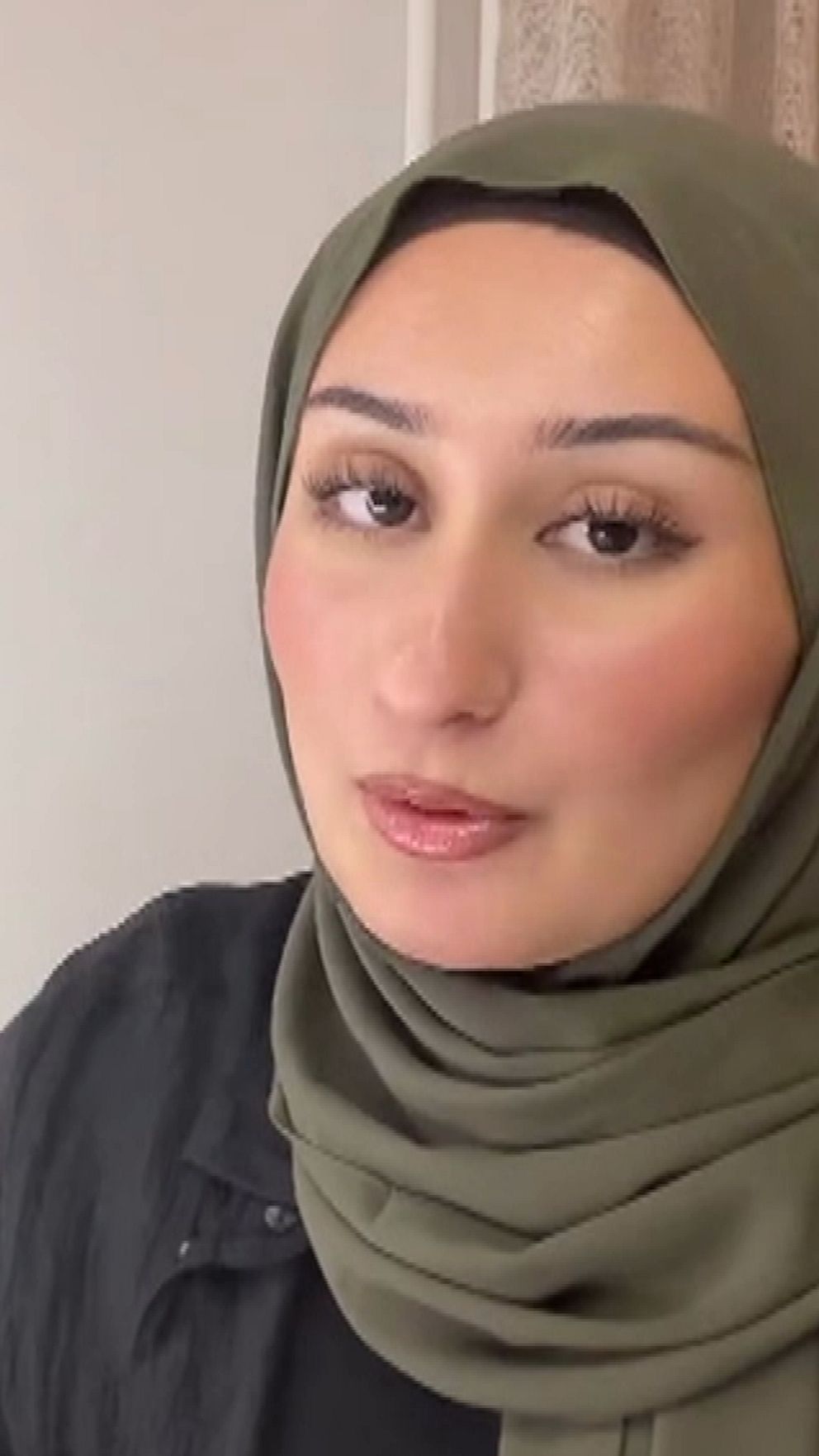 En person i en mörk klädsel och en grön hijab sitter vid ett bord i en inomhusmiljö. I bakgrunden syns en ljus vägg och gardiner, vilket skapar en varm atmosfär.