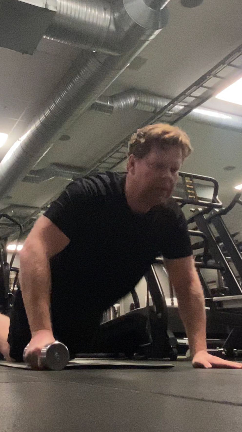 En person utför en träning på golvet i ett gym, med en hantel i handen. Rummet är utrustat med flera träningsmaskiner i bakgrunden.