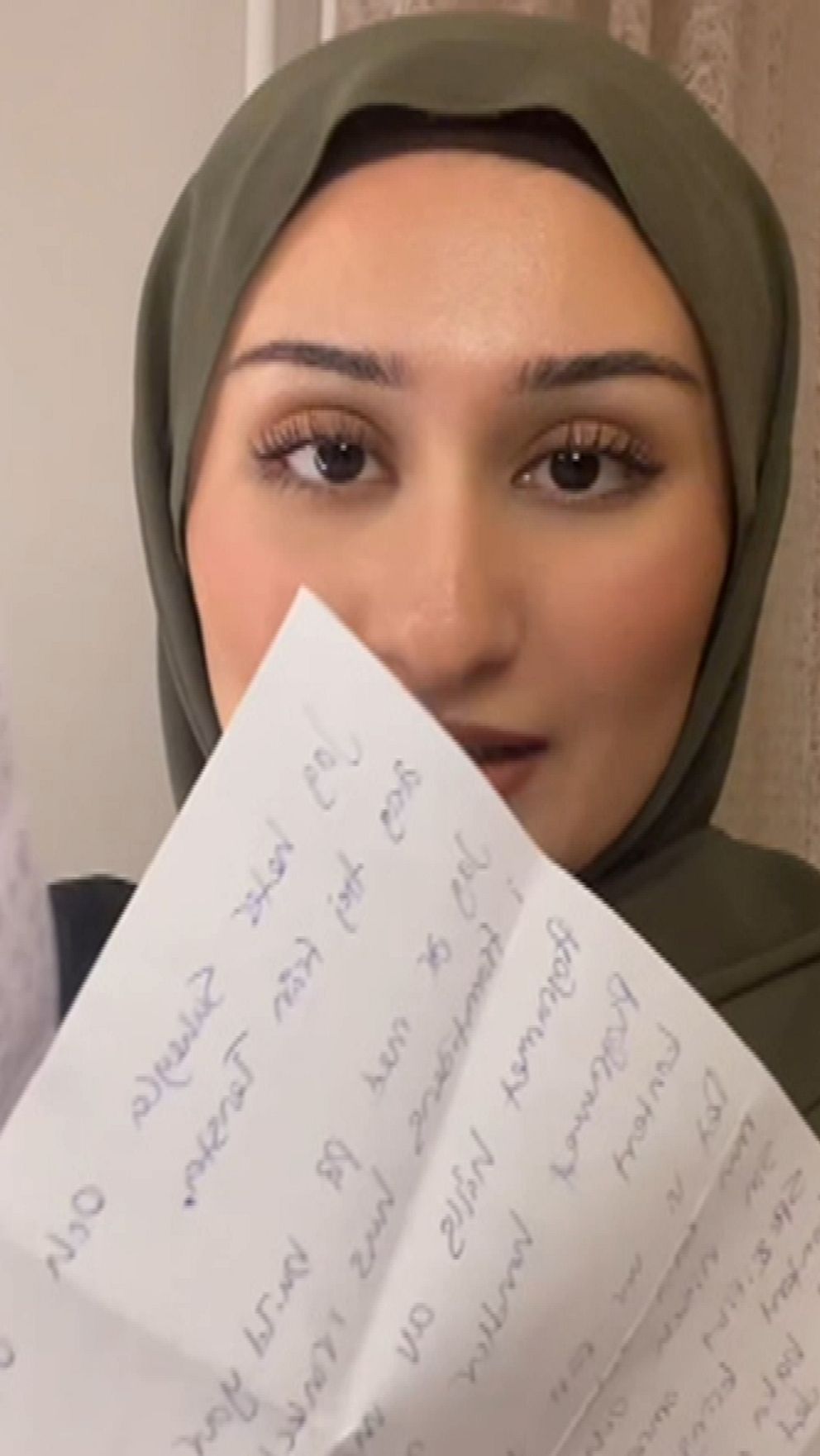 En person som håller upp flera handskrivna papper med text, iklädd en grön huvudduk och med en neutral bakgrund.