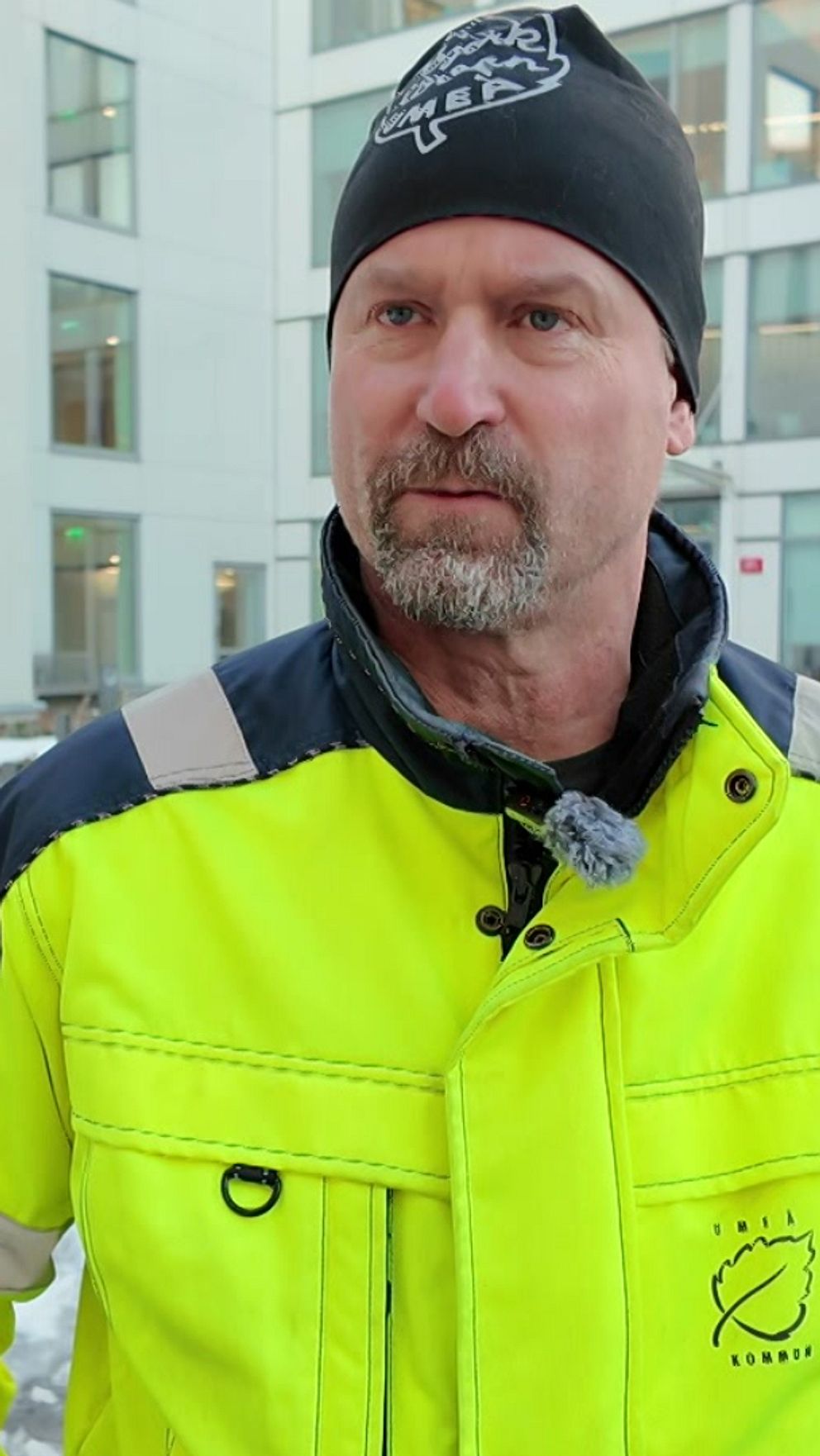 En person i en gul reflexjacka står framför en modern byggnad med snö på marken. I bakgrunden syns flera cyklar parkerade.