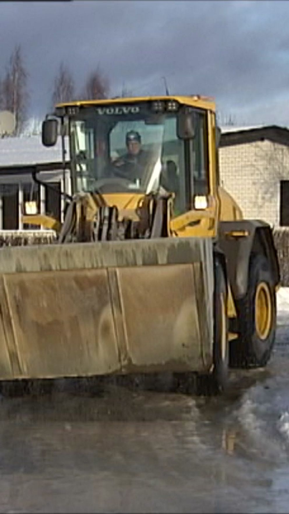 En hjullastare plogar snö på en isig väg framför moderna villor. Himlen är delvis molnig och det finns snö kvar längs vägen och på markerade ytor.