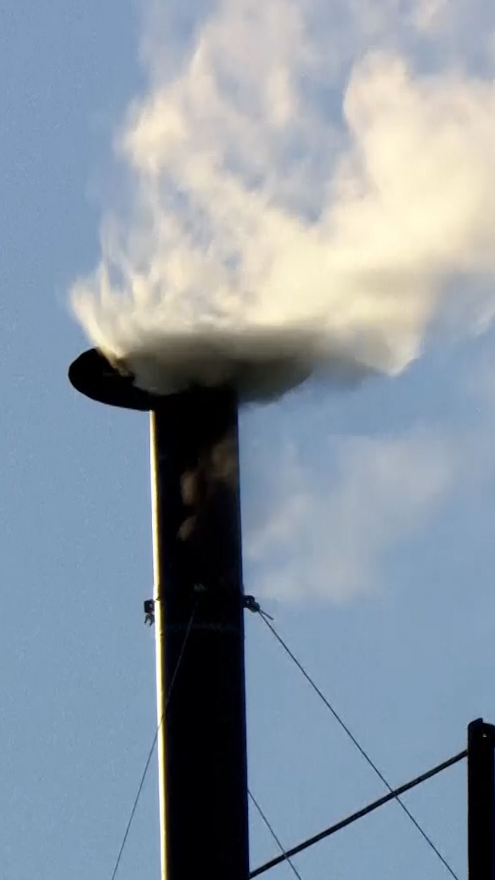 Svart skorsten släpper ut vit rök mot blå himmel.