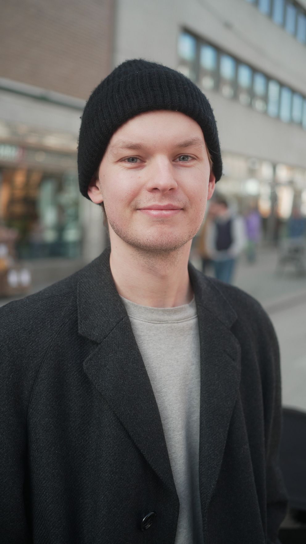 Person med svart mössa och grå kappa står vid en byggnad med skyltfönster.