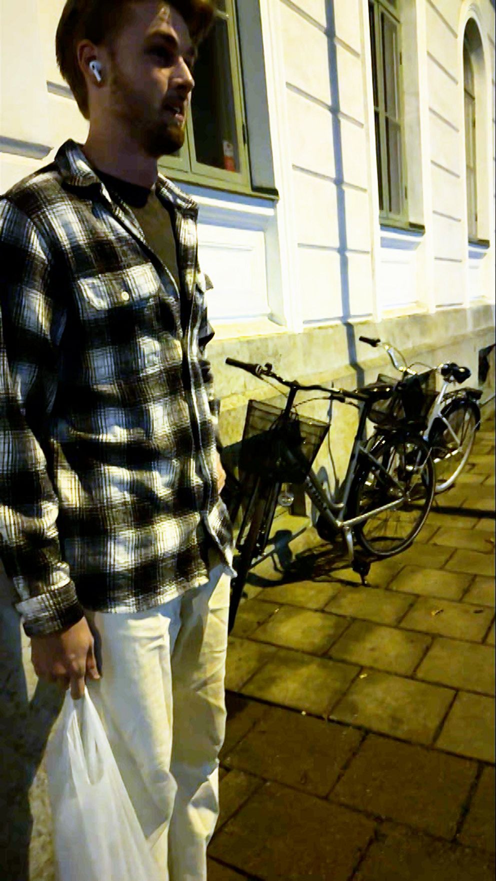 En person står på en trottoar i kvällsbelysning, iförd en rutiga skjorta och vita byxor, medan de håller en plastpåse i ena handen. I bakgrunden syns en cykel parkerad intill en byggnad med ljusa väggar.