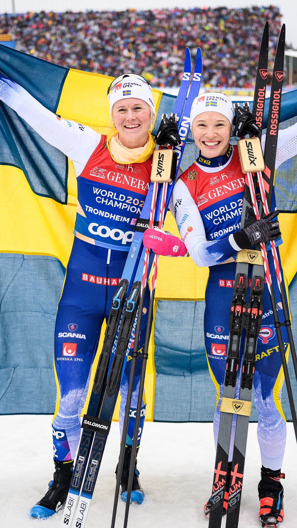 Två svenska längdskidåkare firar med svenska flaggan på en tävling.