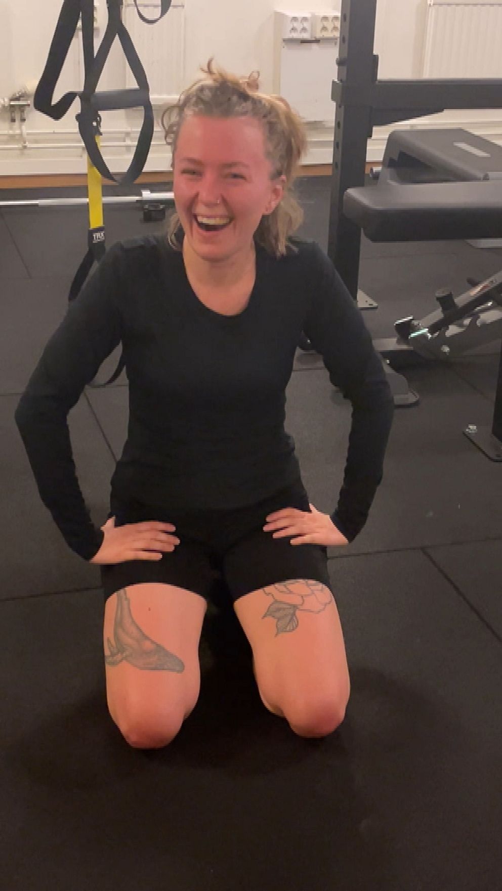 En person sitter på knä på en träningsyta i ett gym med svart gummimatta. Runt omkring finns gymutrustning som TRX-band och en bänk. Personen har en sportig klädsel och ser ut att återhämta sig efter ett träningspass.