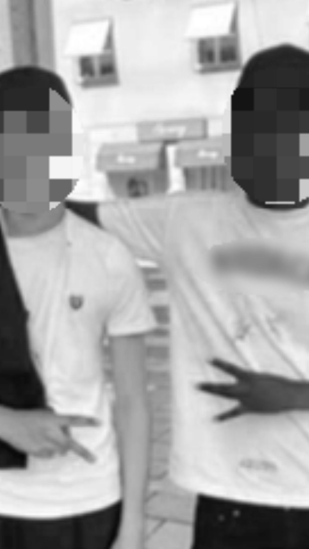 Två personer står tillsammans utanför en byggnad. De ler och poserar för kameran, den ena med armen runt den andra. Båda bär t-shirts, och deras ansikten är pixeliserade för att skydda deras identitet. I bakgrunden syns en stadsmiljö med butiker och en väg.