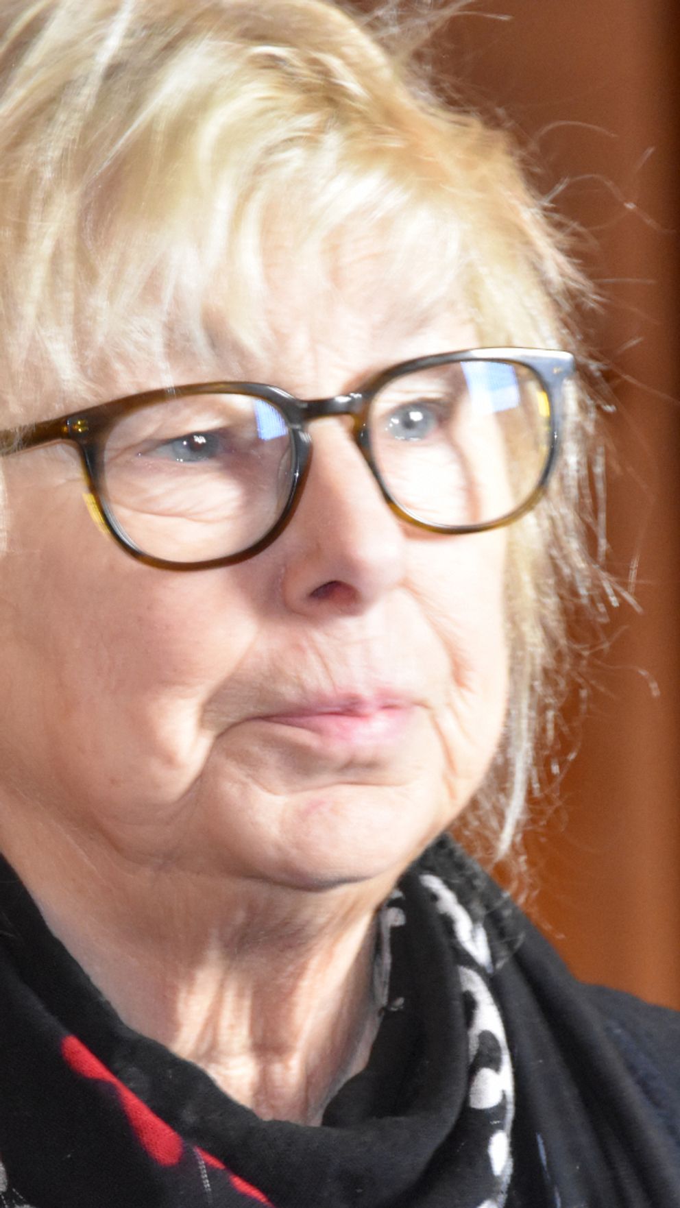 Vänsterpartisten Marita Johansson i en svart jacka och en mönstrad scarf står framför en träpanel.