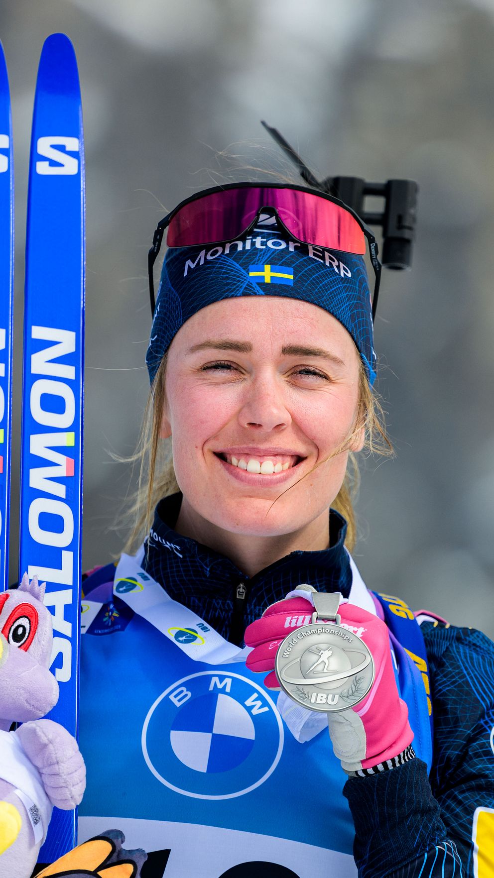 En idrottare klädd i tävlingsdräkt håller upp ett par blå Salomon-skidor och en medalj i ena handen och en lila gosedjursfågel i den andra. Skidgrenar är synliga i bakgrunden.