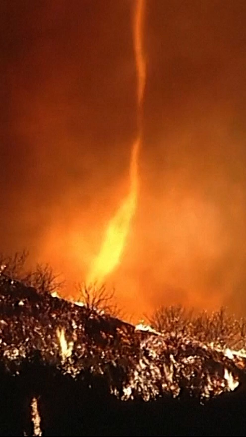 En kraftig eldsvåda tillsammans med en eldtornado rasar över ett bergslandskap, med lågor som slår upp mot himlen och en eldvirvel som transporterar rök och gnistor.