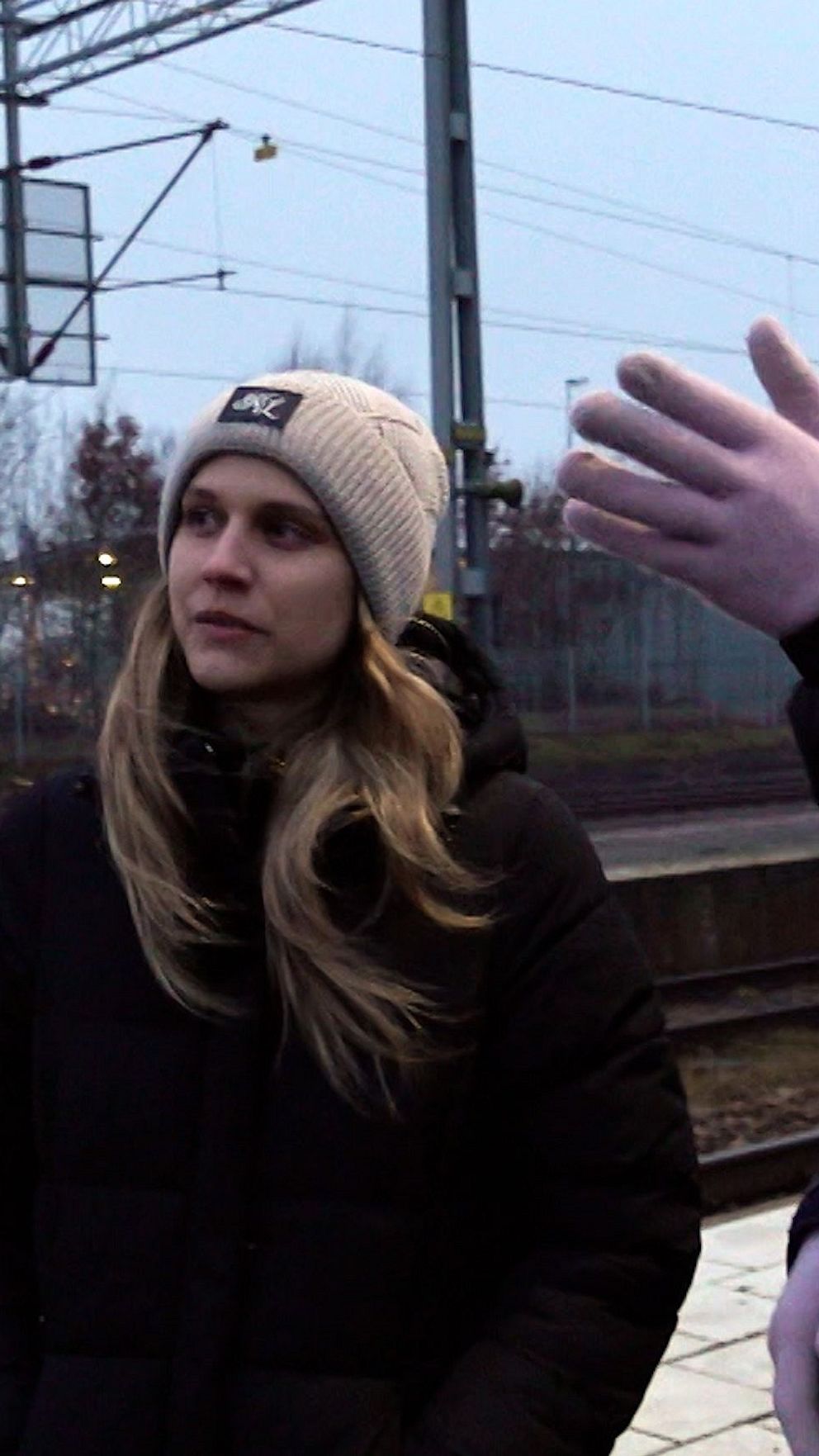 Person i mörk jacka och mössa på tågperrong, bredvid syns en uppsträckt hand i förgrunden.
