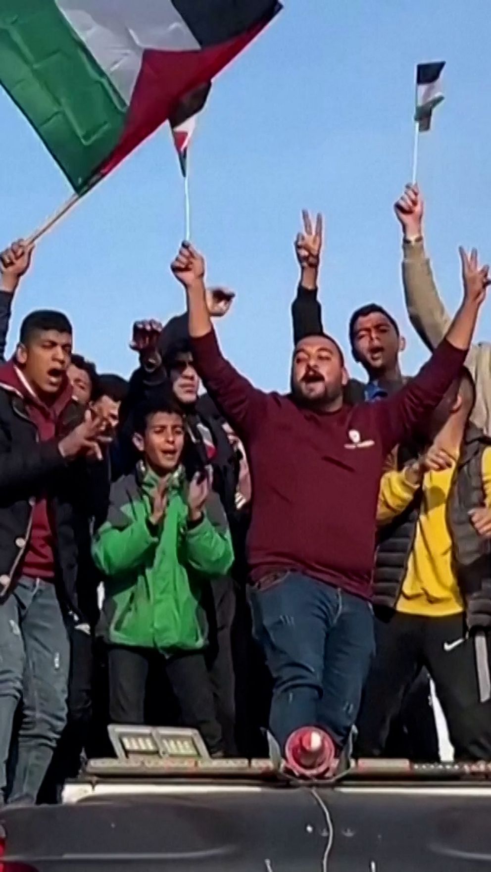 Människor firar och demonstrerar på taket av en bil, håller upp palestinska flaggor och gör segervisslande gester. Bakgrunden består av byggnader i en stadsmiljö.