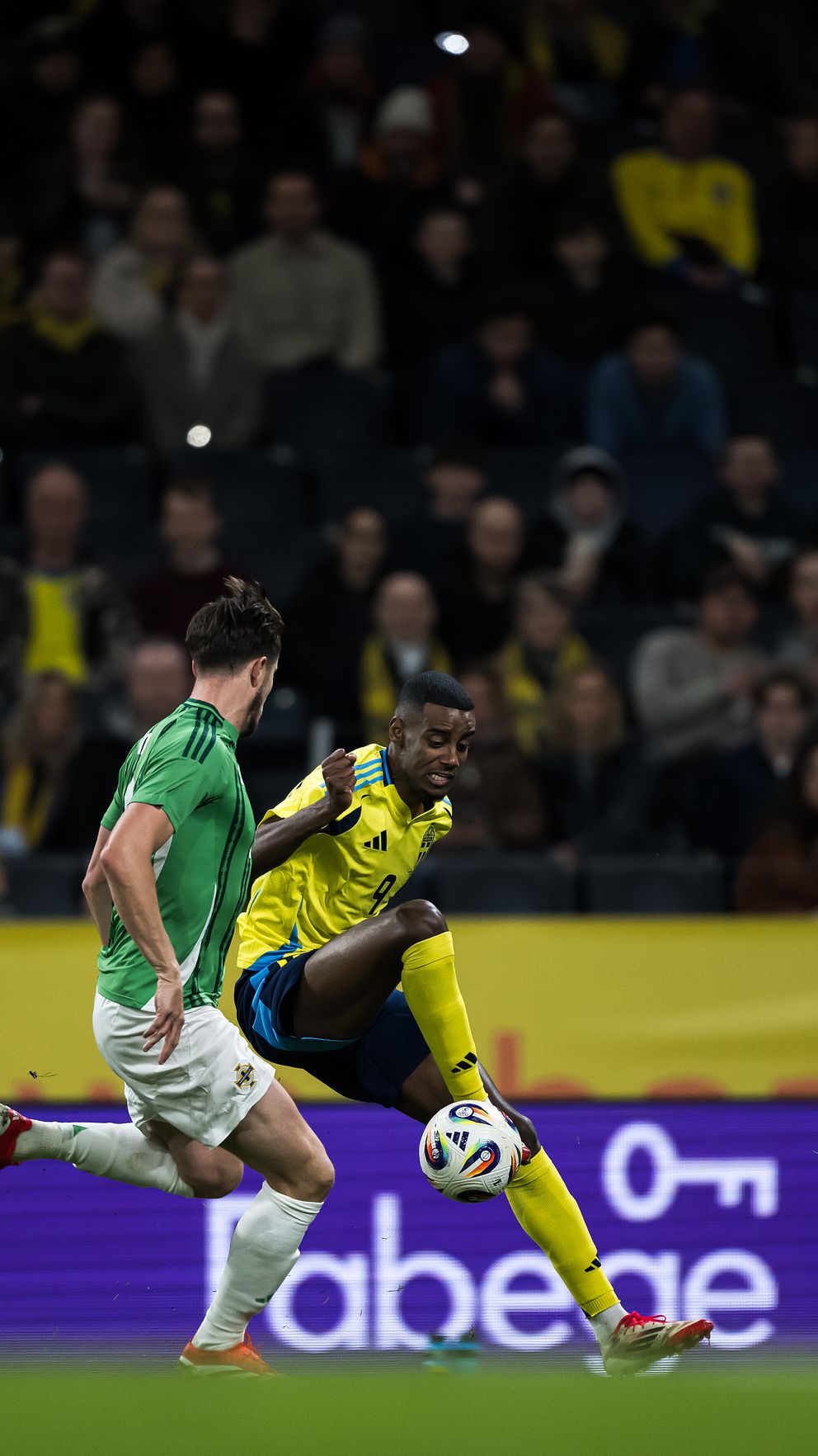 Fotbollsspelare i grön och gul tröja tävlar om bollen på planen.
