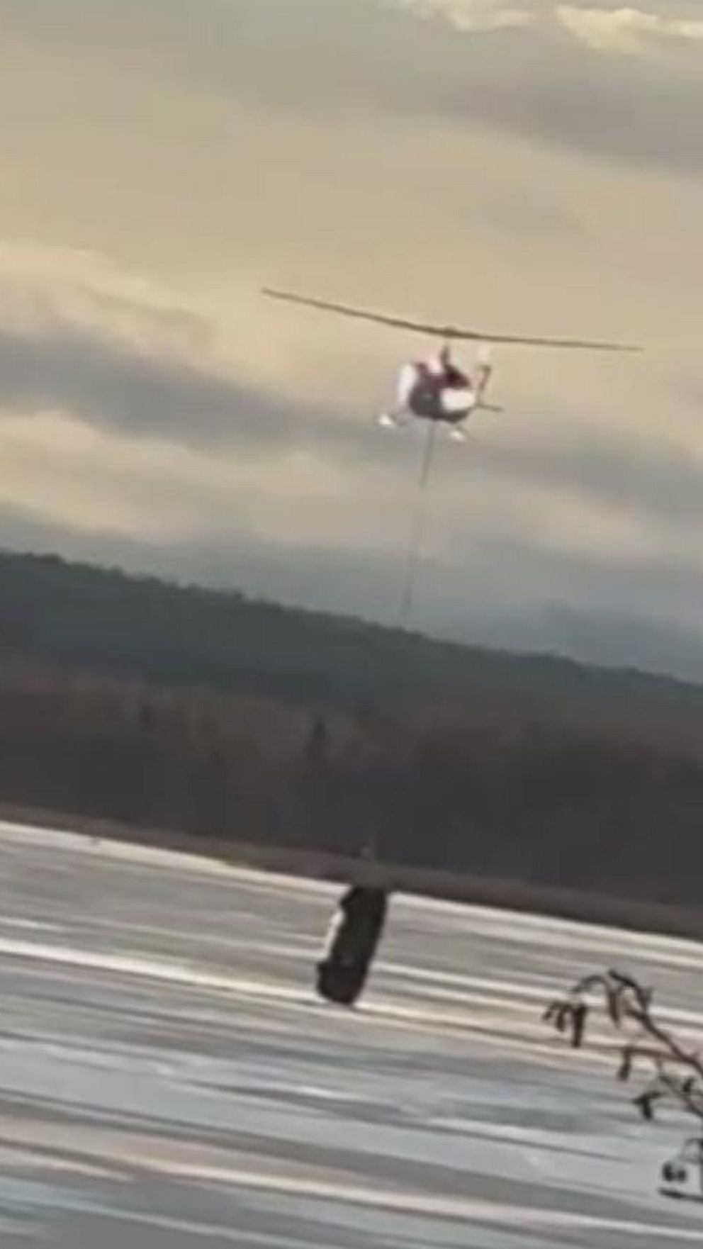 En helikopter flyger över ett fruset landskap och bär en bil under sig. I bakgrunden syns skogsklädda kullar och en molntäckt himmel.