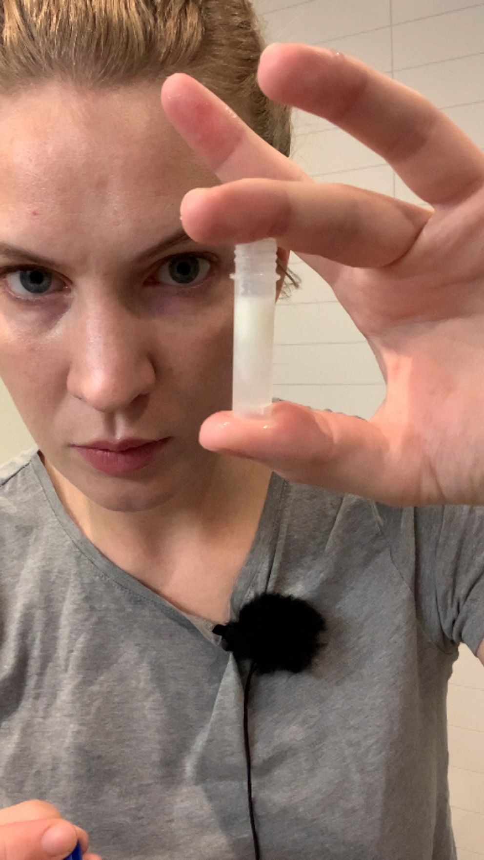 Person i grå tröja håller upp ett provrör med spott framför sig, mikrofon syns på klädsel.