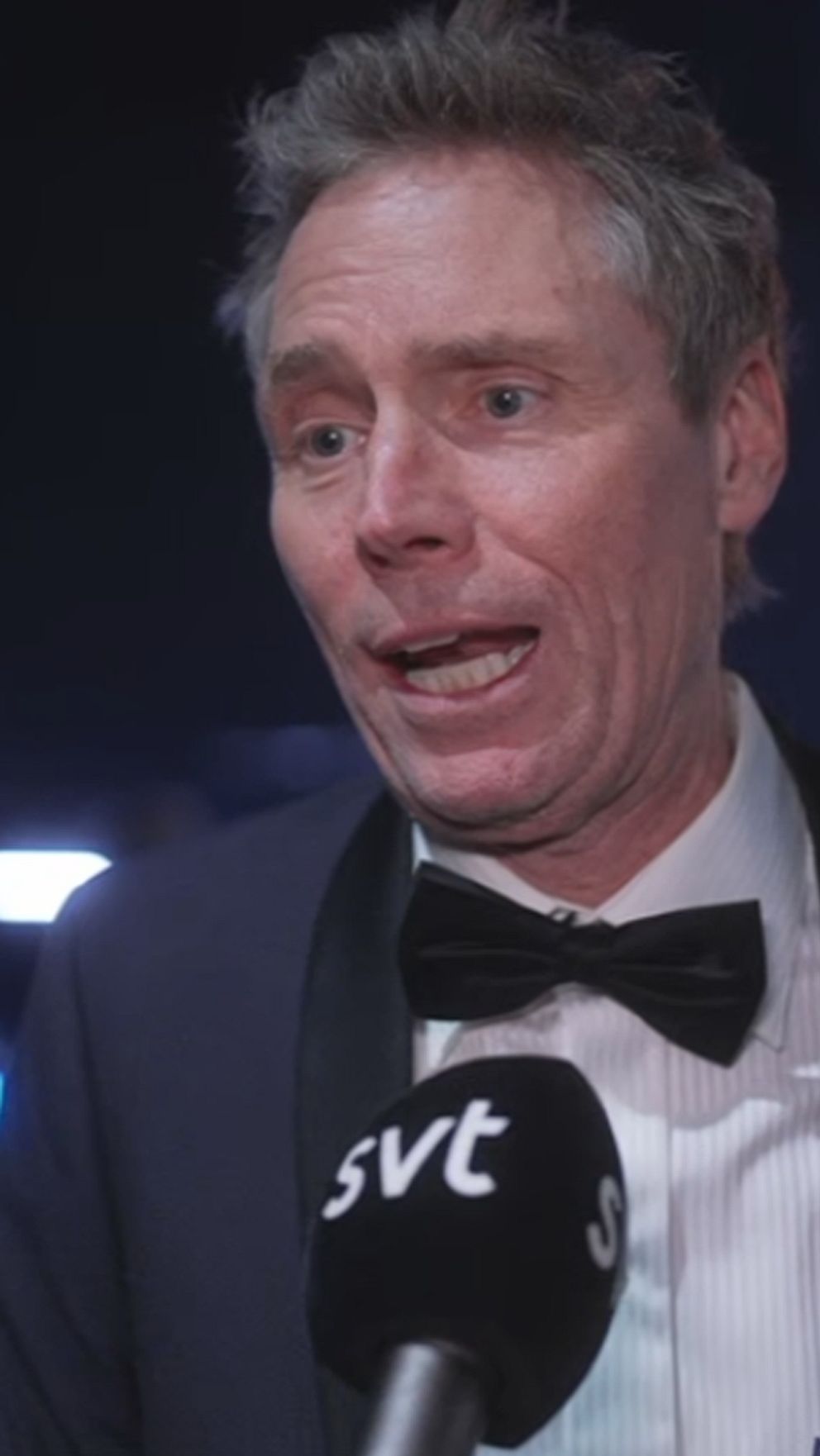En man i kostym med fluga intervjuas, stående i en scenisk miljö med blå belysning. Bakgrunden visar suddiga konturer av människor och ljus, vilket tyder på ett evenemang eller en gala.