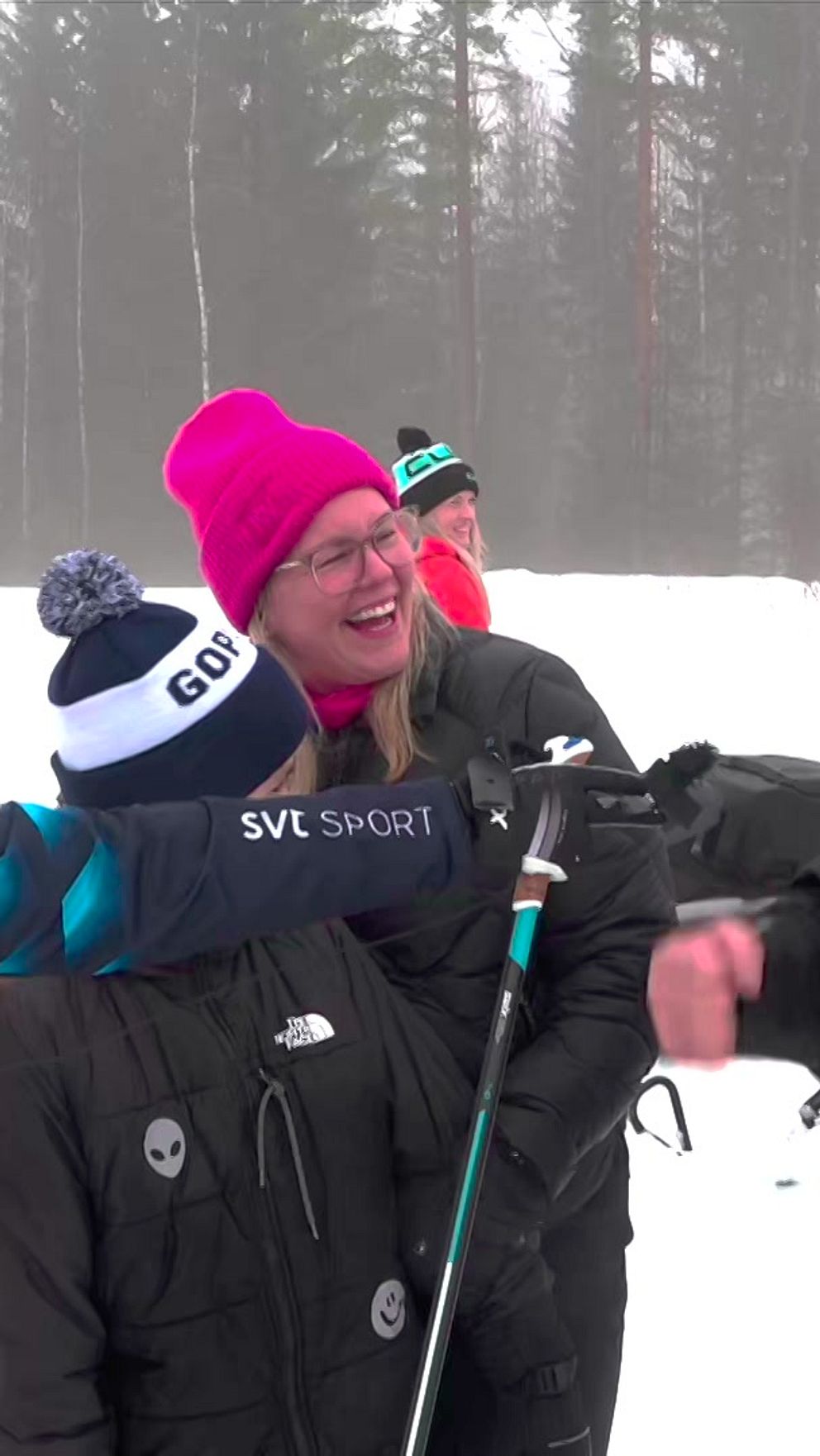 En grupp människor skrattar utomhus i vinterkläder med snö och träd i bakgrunden.